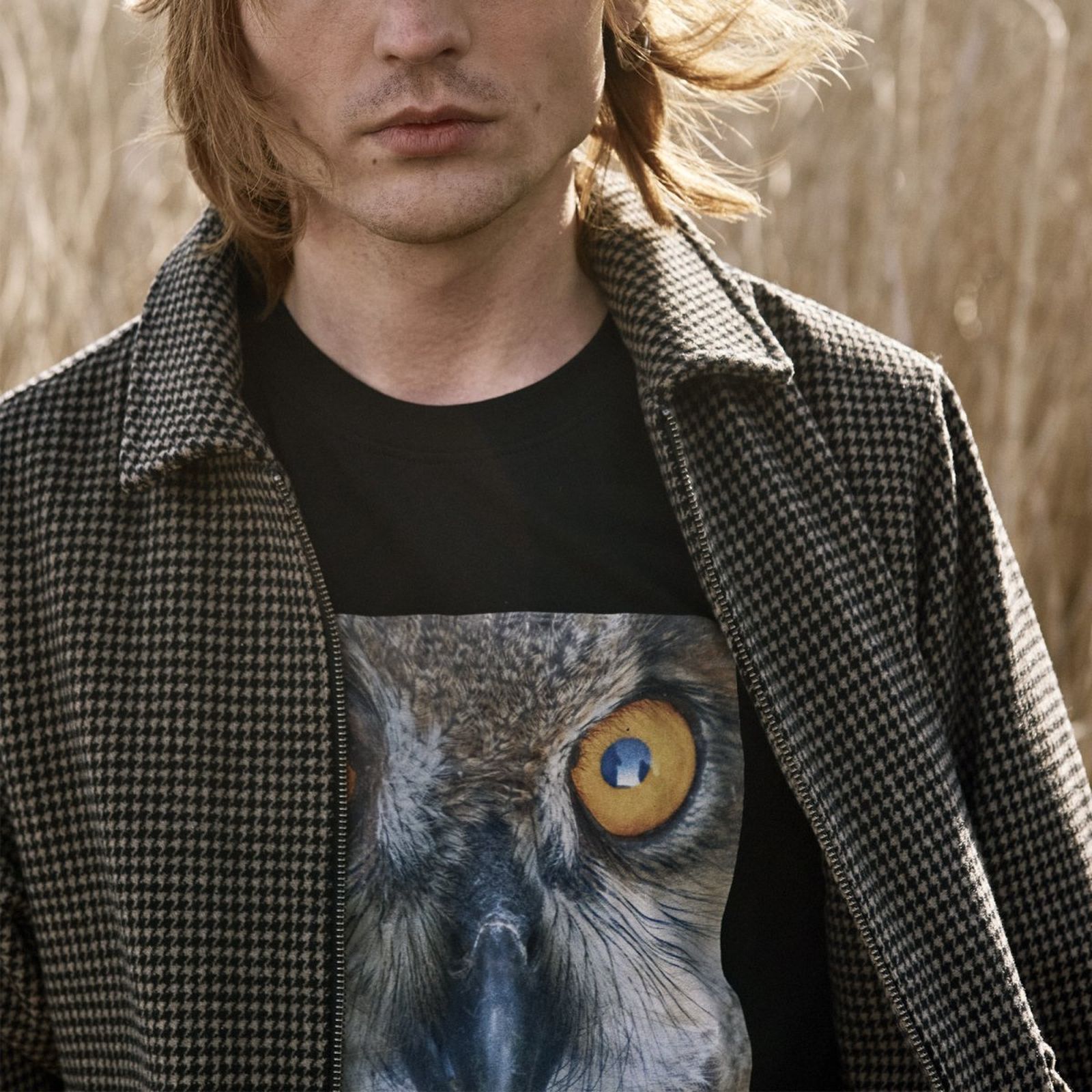 Person in a checkered jacket stands outdoors, wearing a black T-shirt with a large owl eye print. Brown, windswept hair partially covers face.