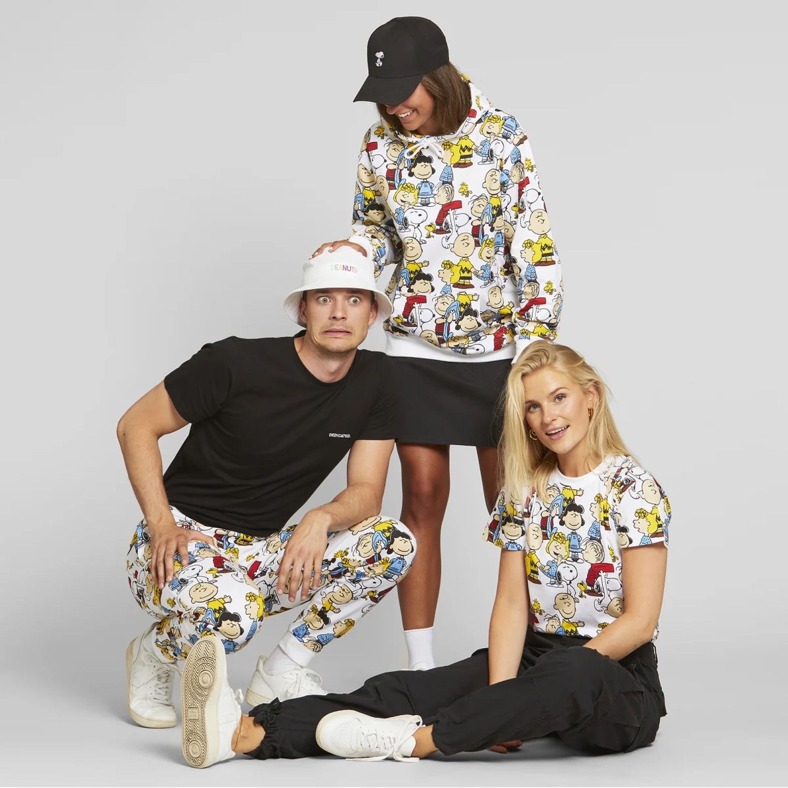 Three people in casual outfits, two wearing matching cartoon-patterned clothes, and one in a black shirt and cap, pose against a gray background.