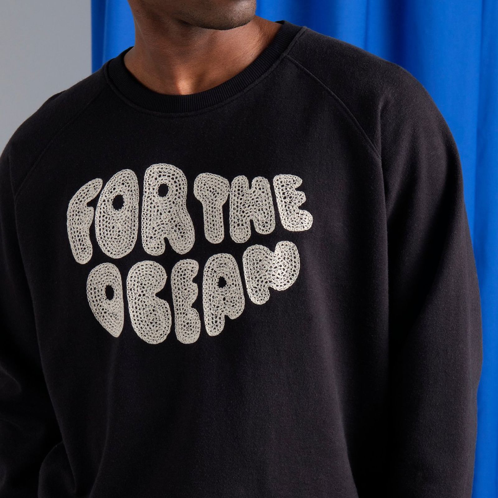 Person wearing a black sweatshirt with "FOR THE OCEAN" in textured white letters, standing against a blue curtain.