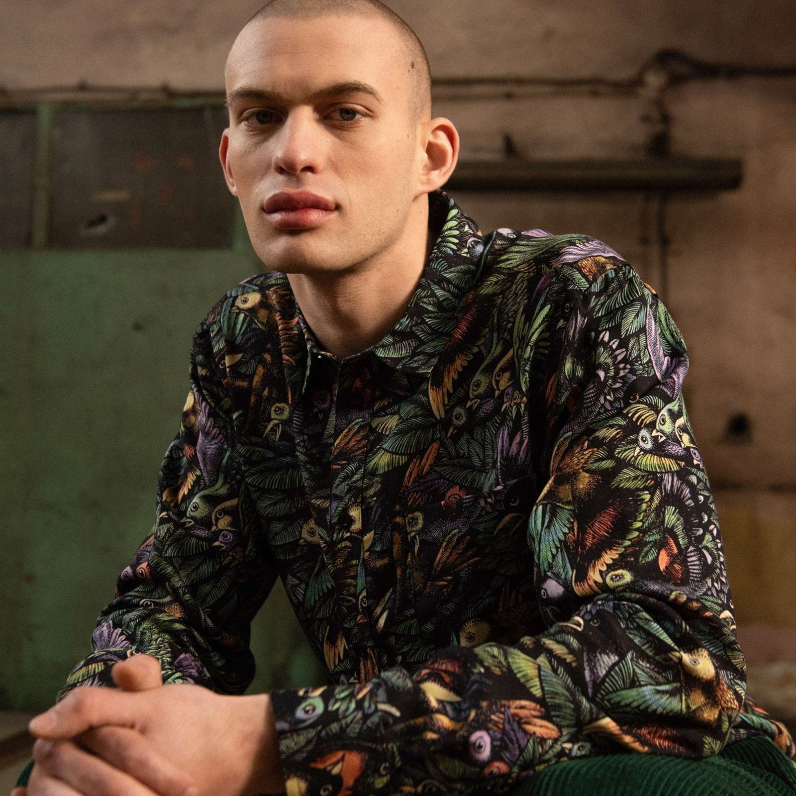 A person with a shaved head wearing a colorful, patterned shirt sits indoors, looking at the camera. The background is industrial.