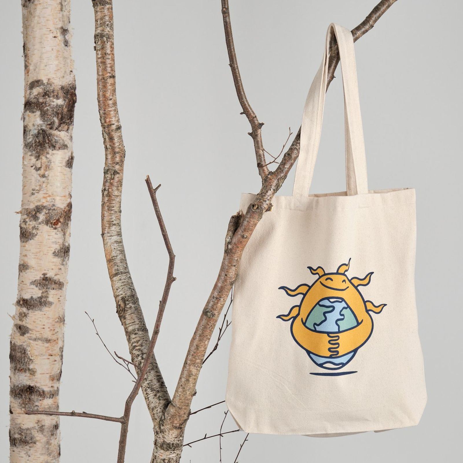 A beige tote bag with a sun and earth design hangs on a tree branch against a plain background.