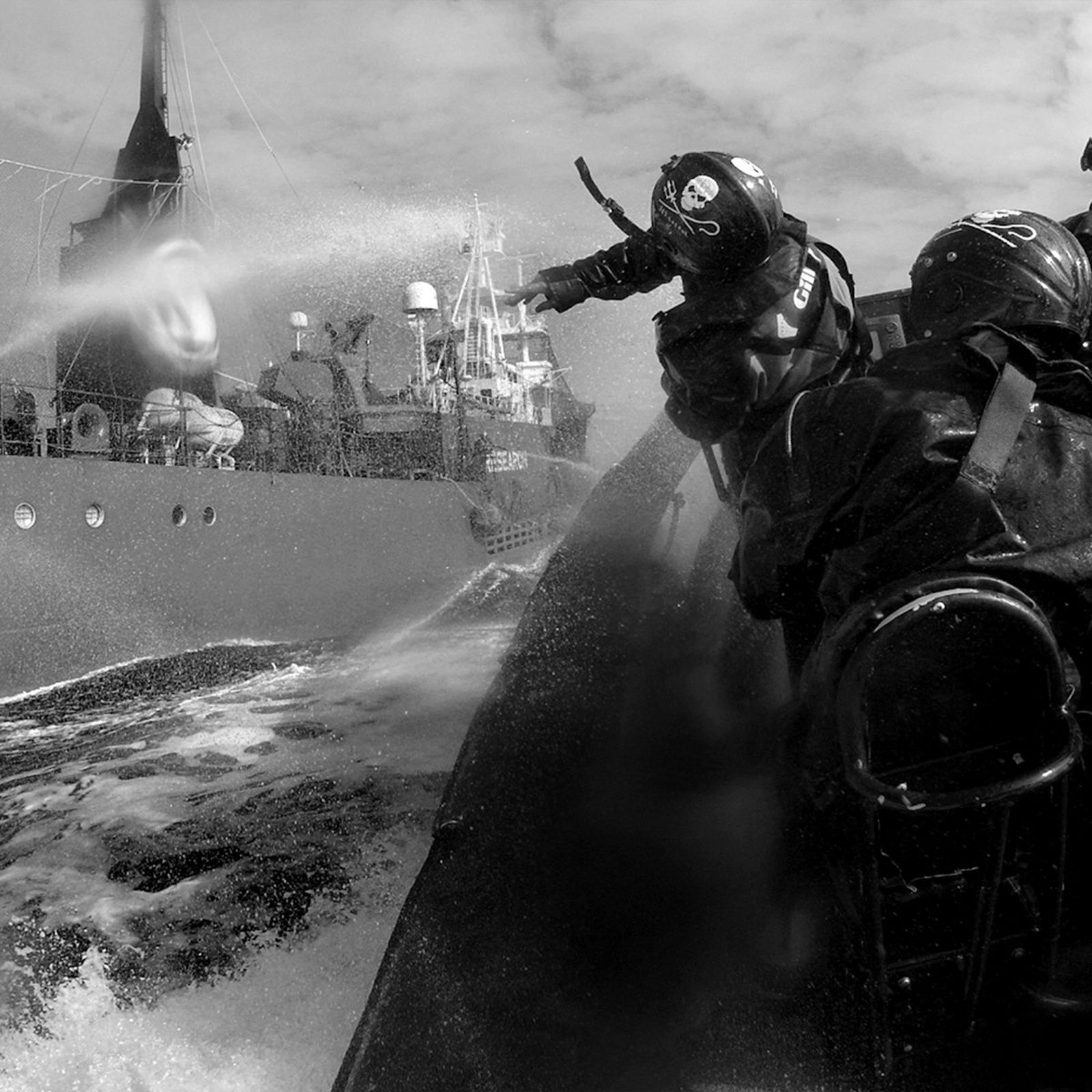 Two people in a boat spray water at a larger ship, creating a dramatic scene at sea. The image is in black and white.