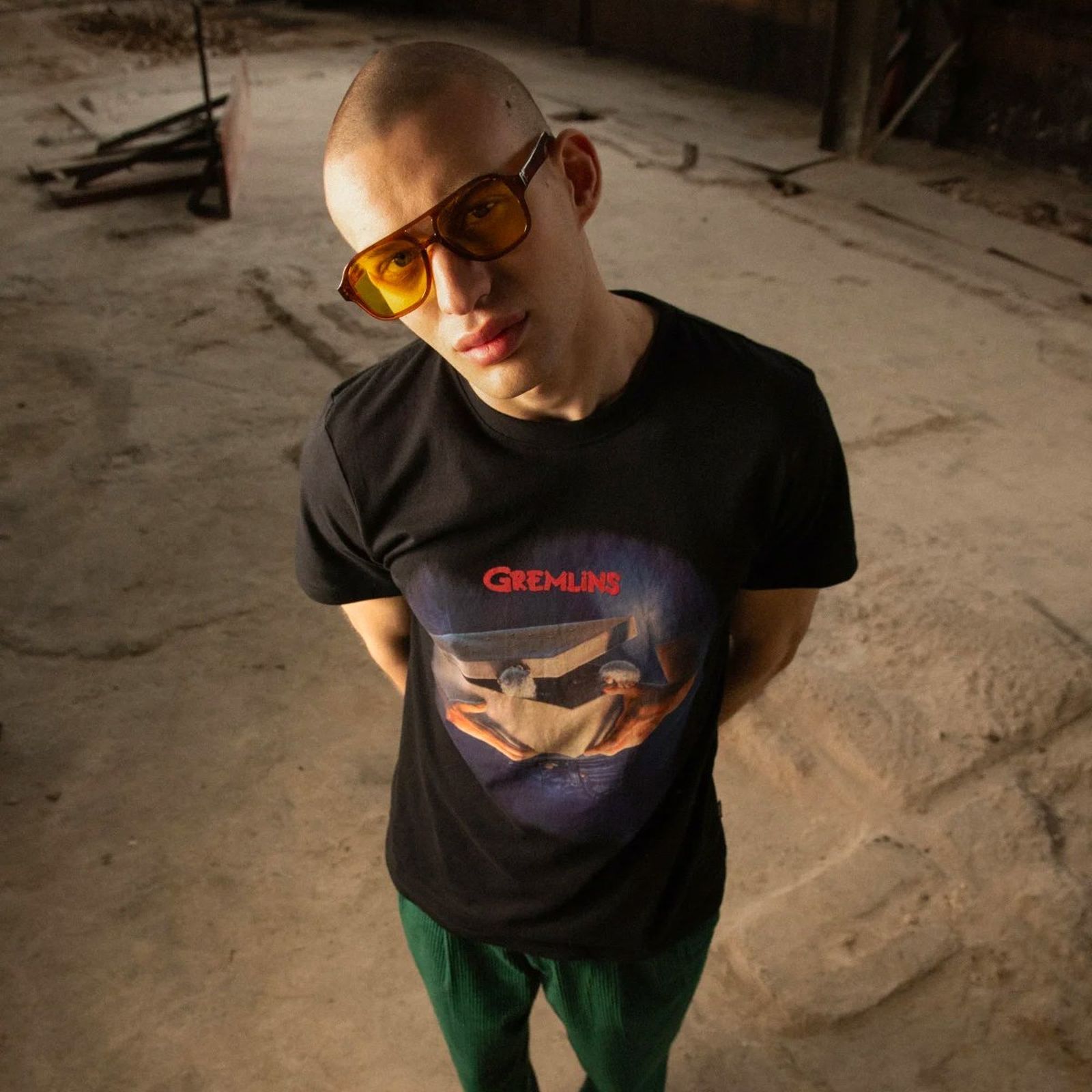 Person wearing sunglasses and a "Gremlins" T-shirt, standing on a dusty floor, looking up at the camera.