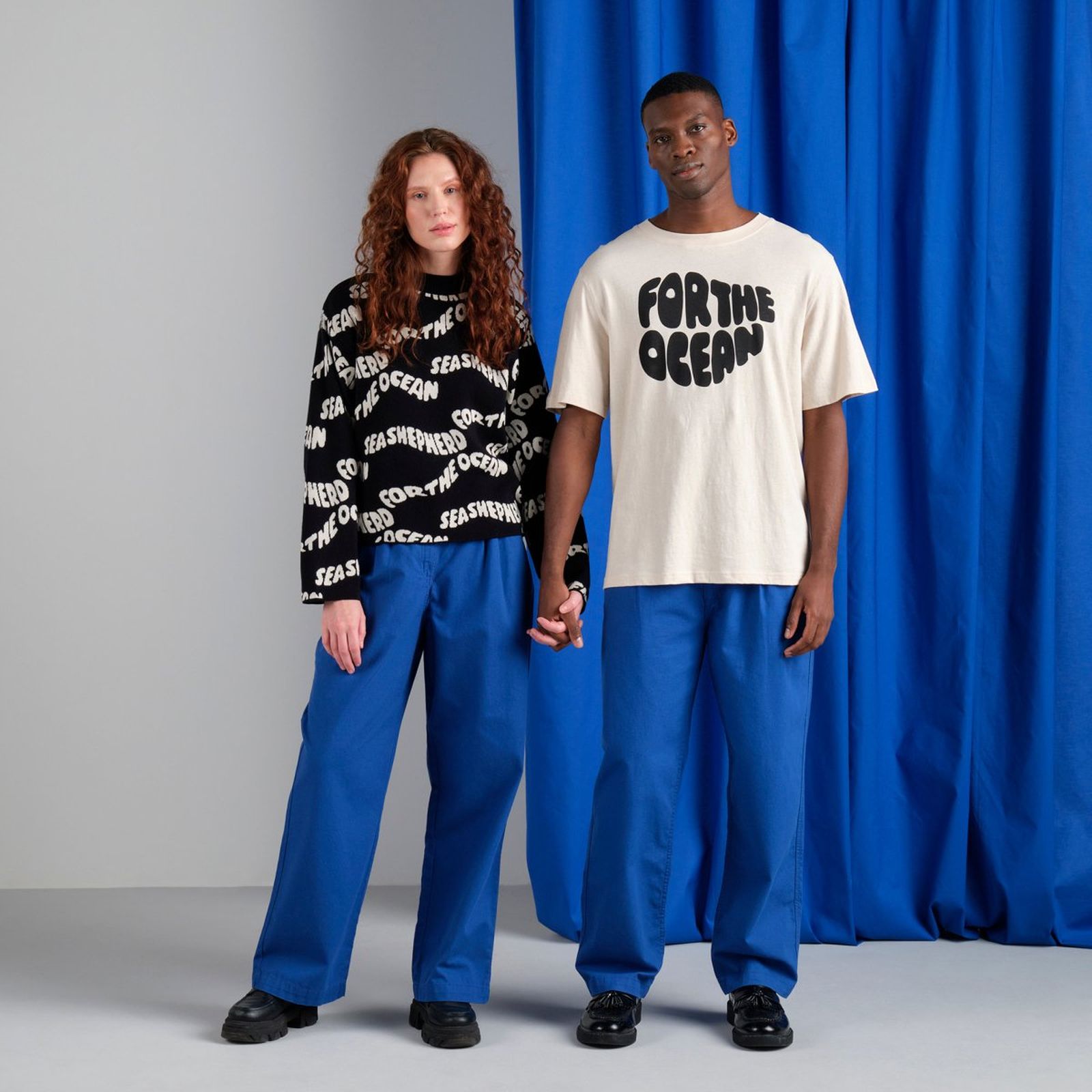 A couple stands holding hands, both wearing blue pants. She has a black "Sea Shepherd" sweater, and he has a white "For the Ocean" T-shirt.
