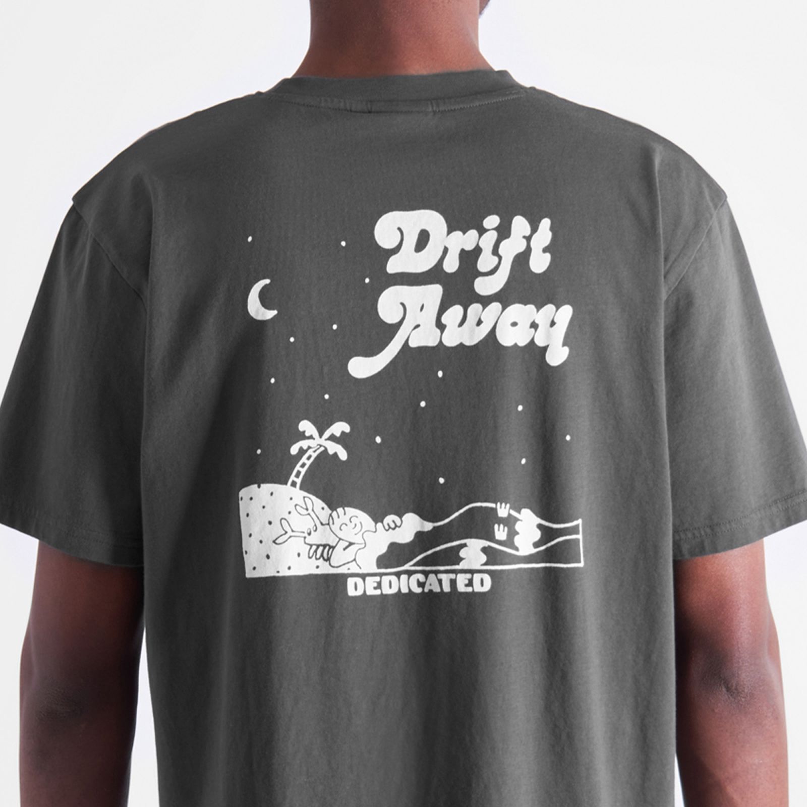 Person wearing a gray T-shirt with a white graphic of a person relaxing on a beach under a crescent moon, text reads "Drift Away Dedicated."