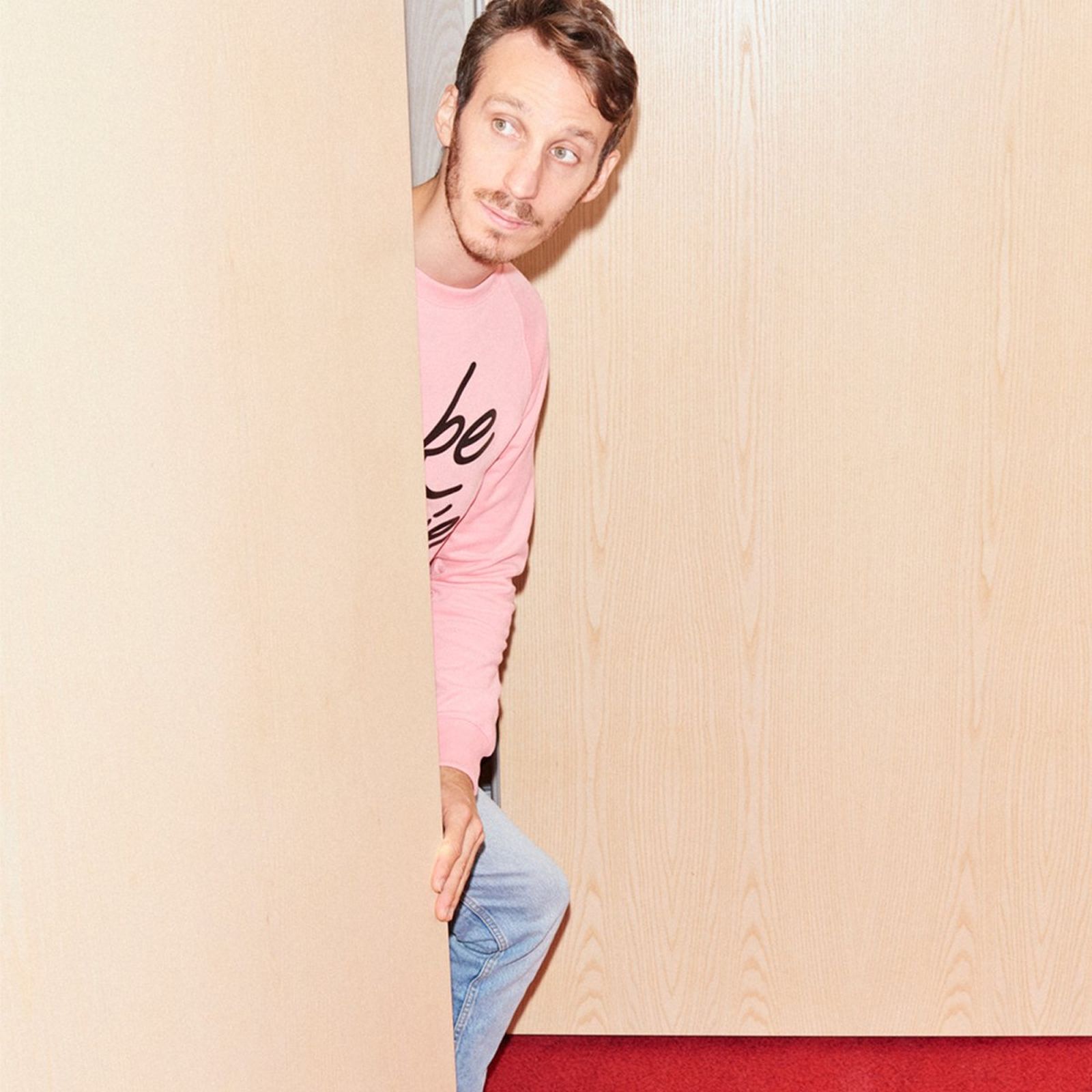 Man in a pink sweater and jeans peeks around a wooden door, standing on a red carpeted floor.