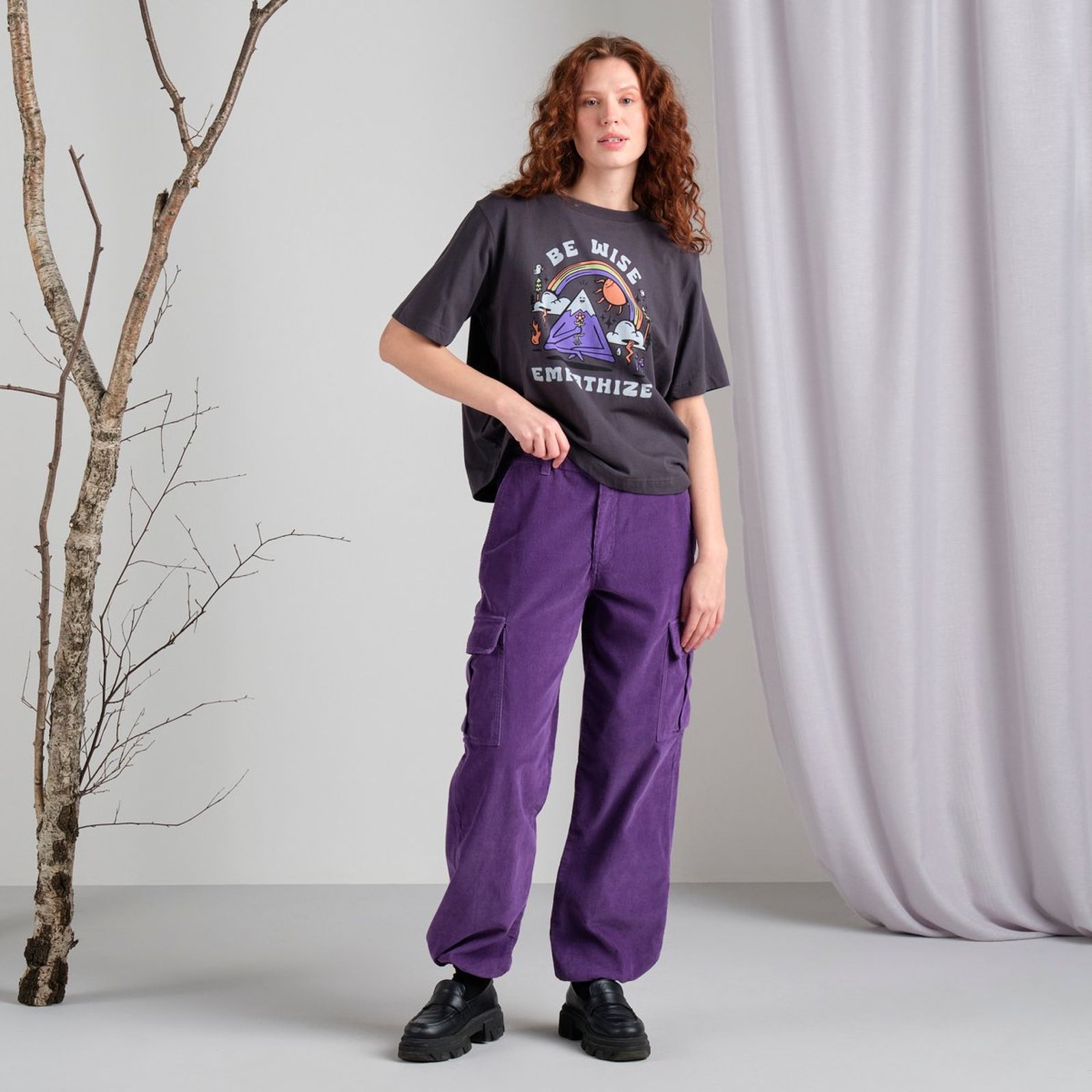 Woman with curly hair wearing a graphic tee and purple cargo pants stands near a bare branch, against a light gray curtain backdrop.