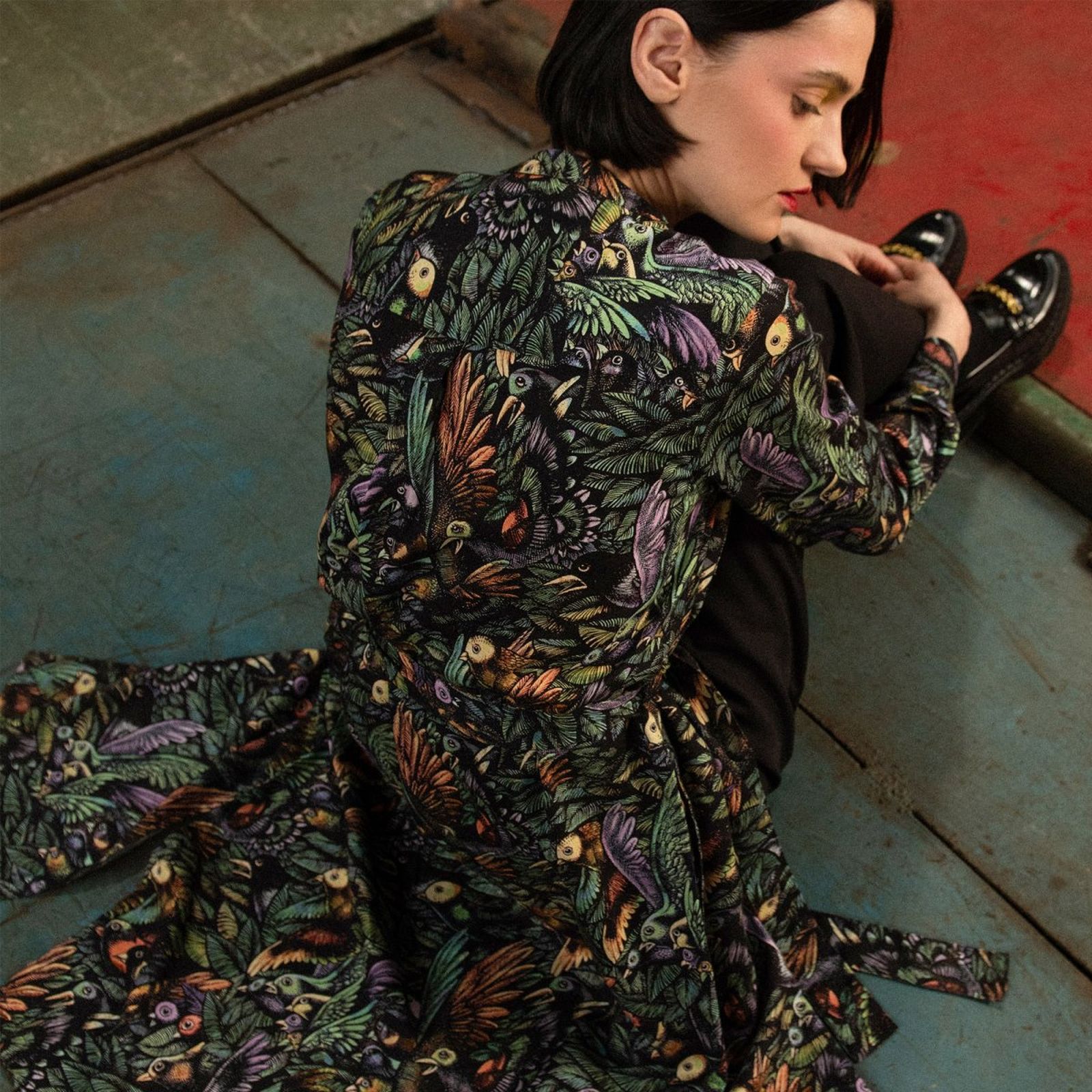 Woman in a colorful, patterned outfit sits on a textured floor with her knees up, looking contemplative.