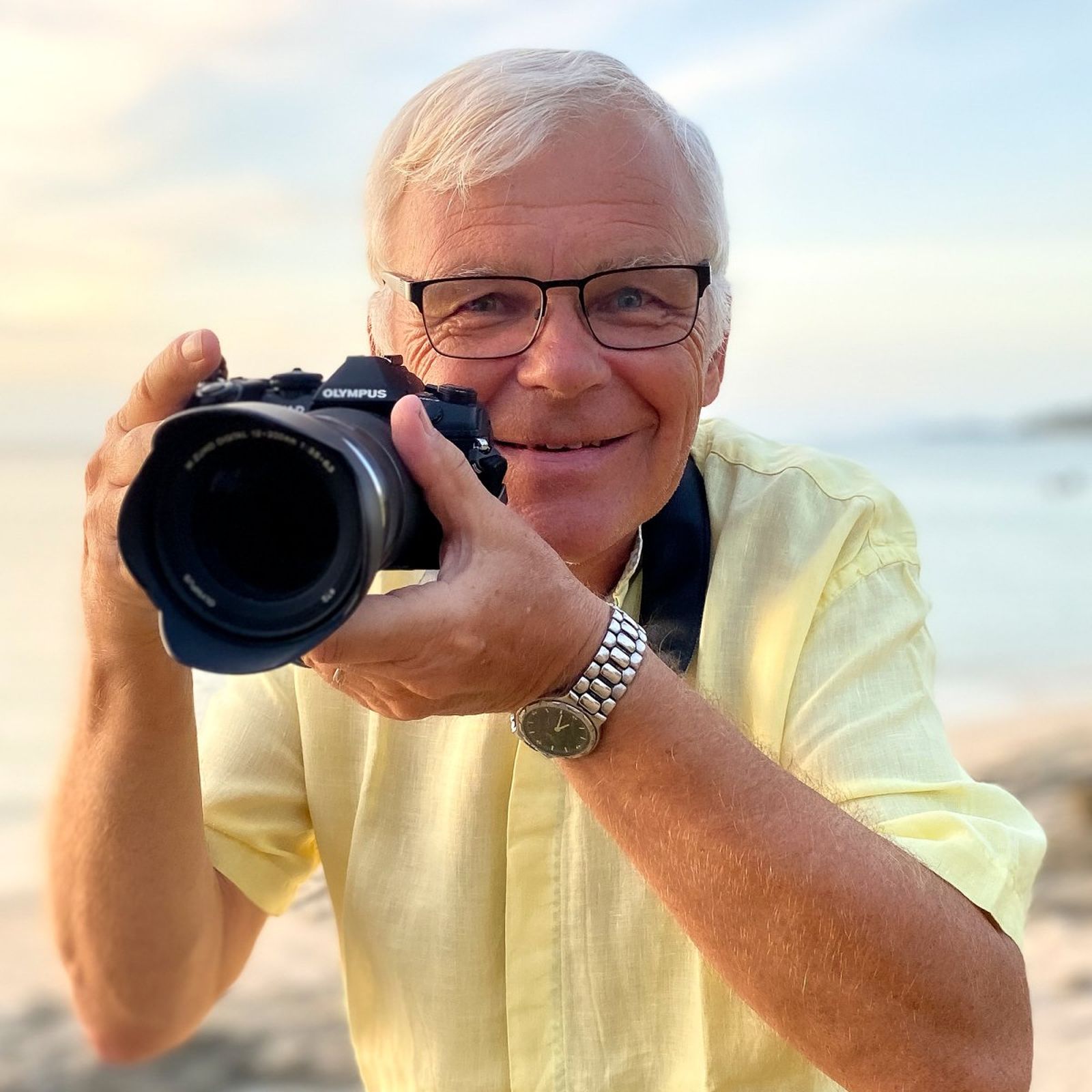Elderly man in a yellow shirt smiling and holding a camera on a beach at sunset.