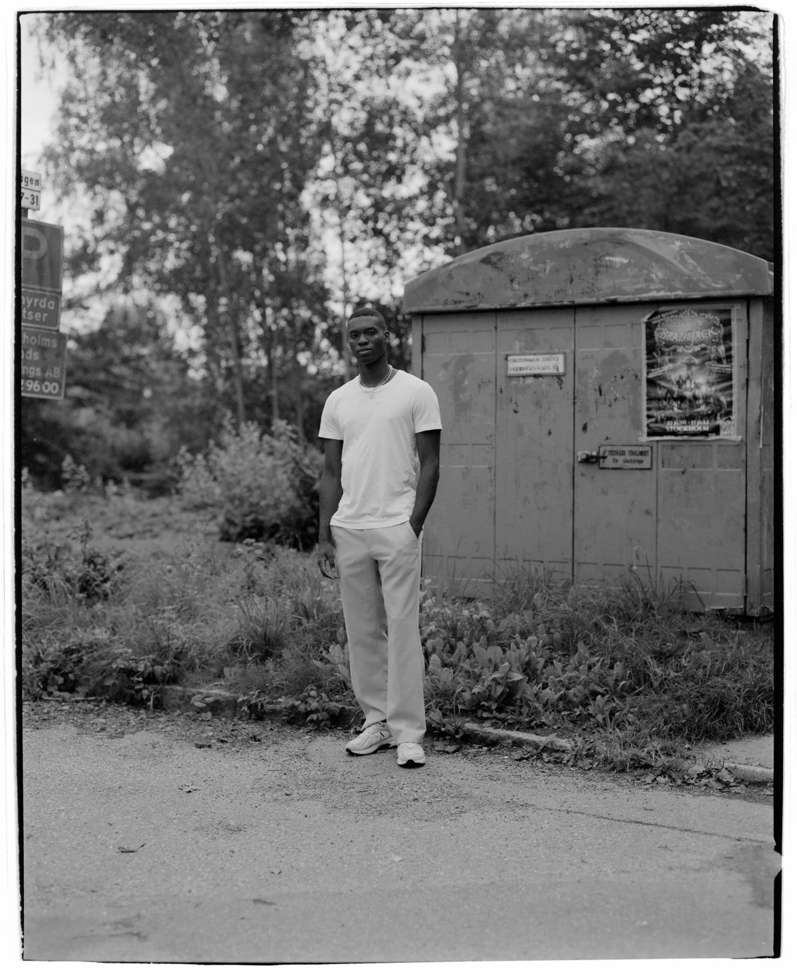 Man in a white t-shirt and pants stands on a quiet street near a weathered shed, surrounded by trees and greenery. Black and white photo.