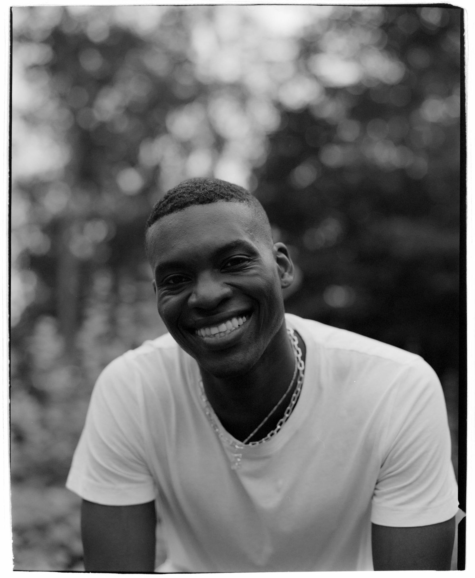 Smiling person in a white shirt and necklace, outdoors with blurred trees in the background, captured in black and white.