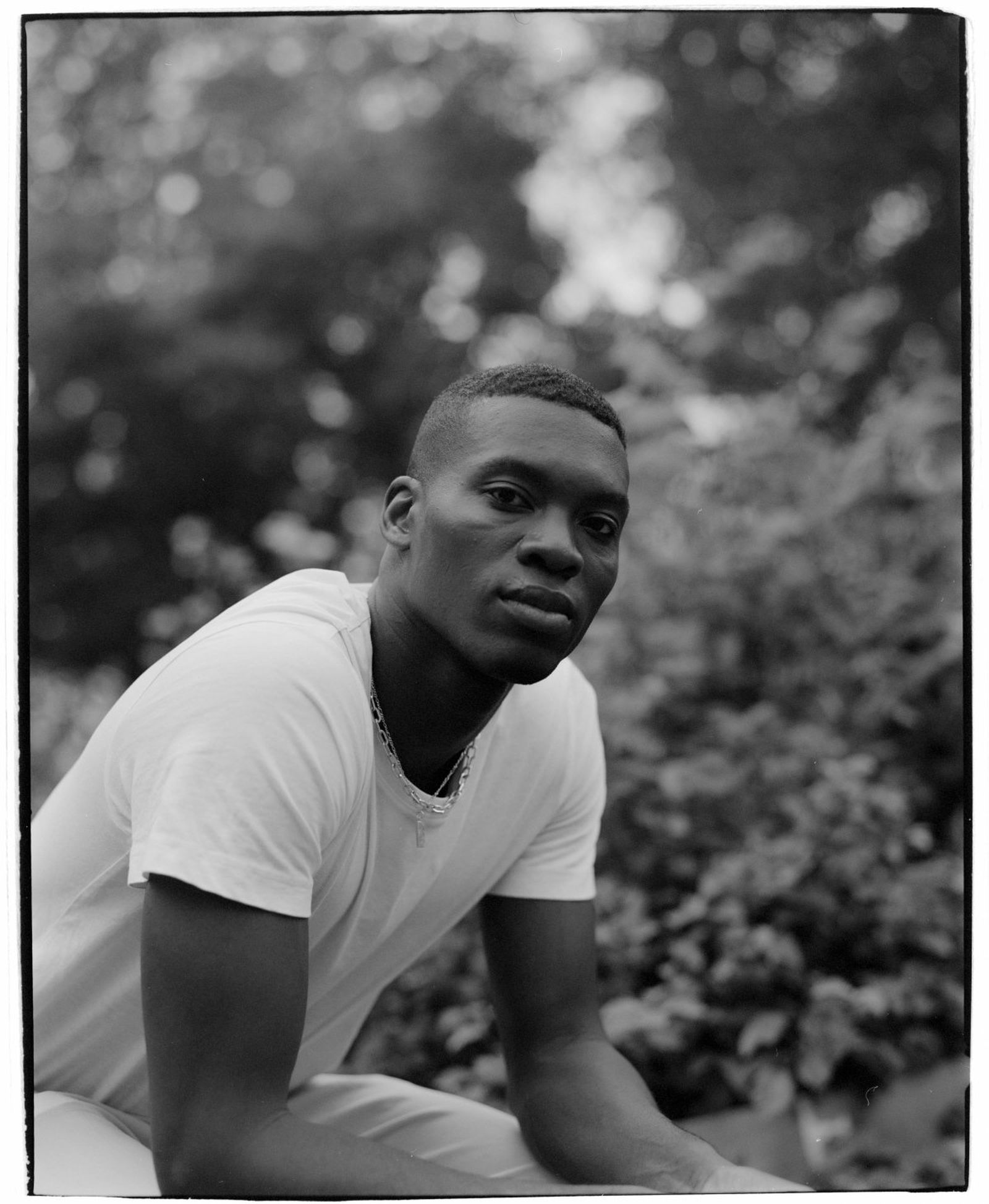 Black and white portrait of a person in a white t-shirt, sitting outdoors with blurred foliage in the background.
