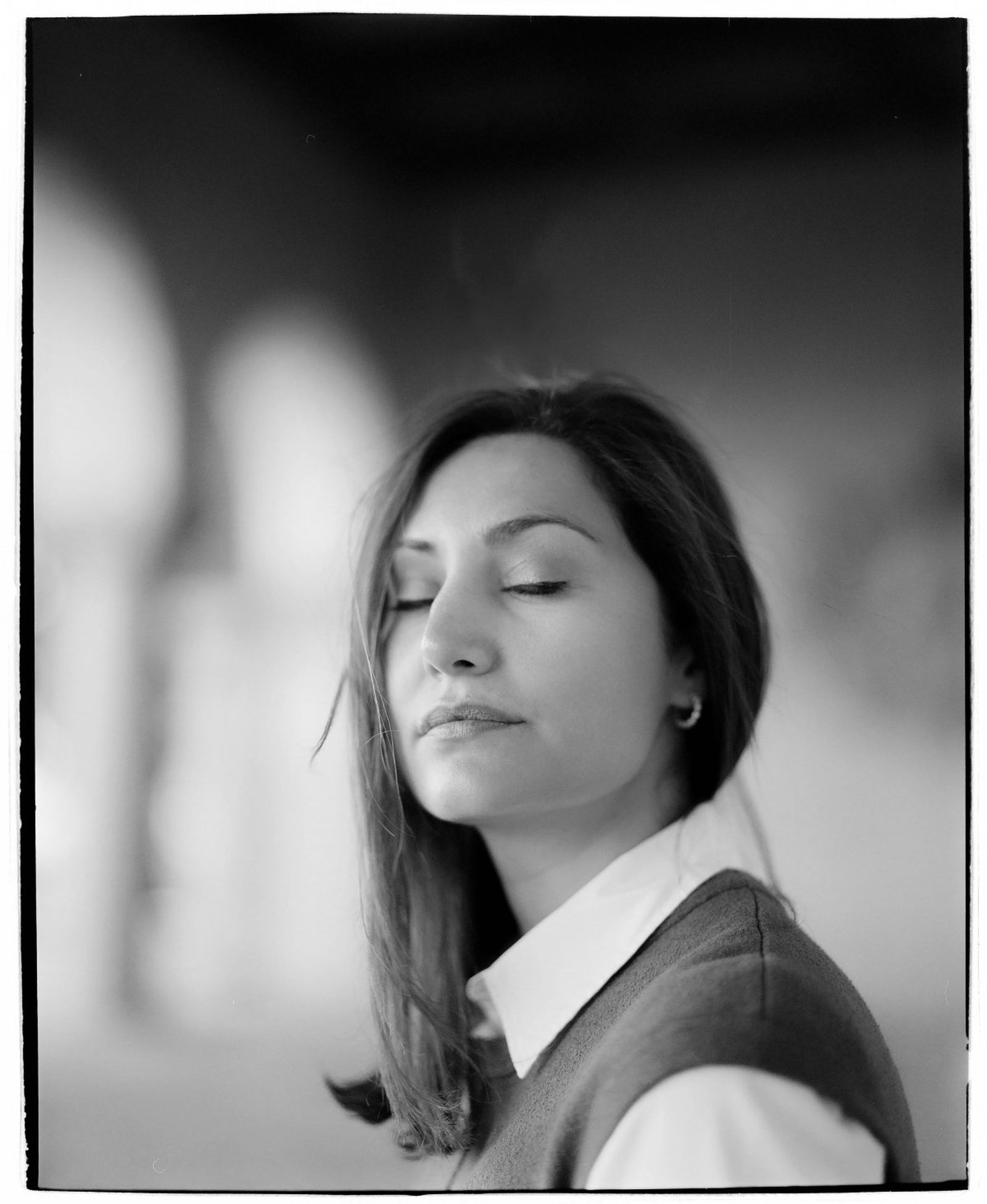 Black and white portrait of a person with long hair, eyes closed, wearing a collared shirt and sweater, in a softly blurred background.