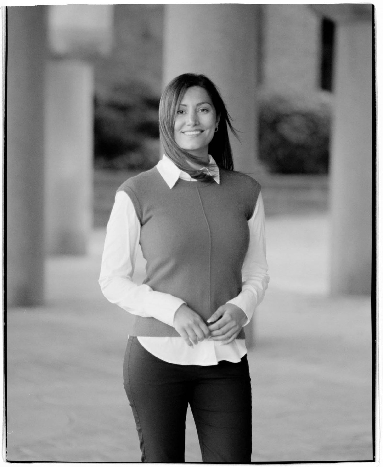A woman smiling, wearing a sweater vest over a collared shirt, standing outdoors with blurred columns and foliage in the background. Black and white.