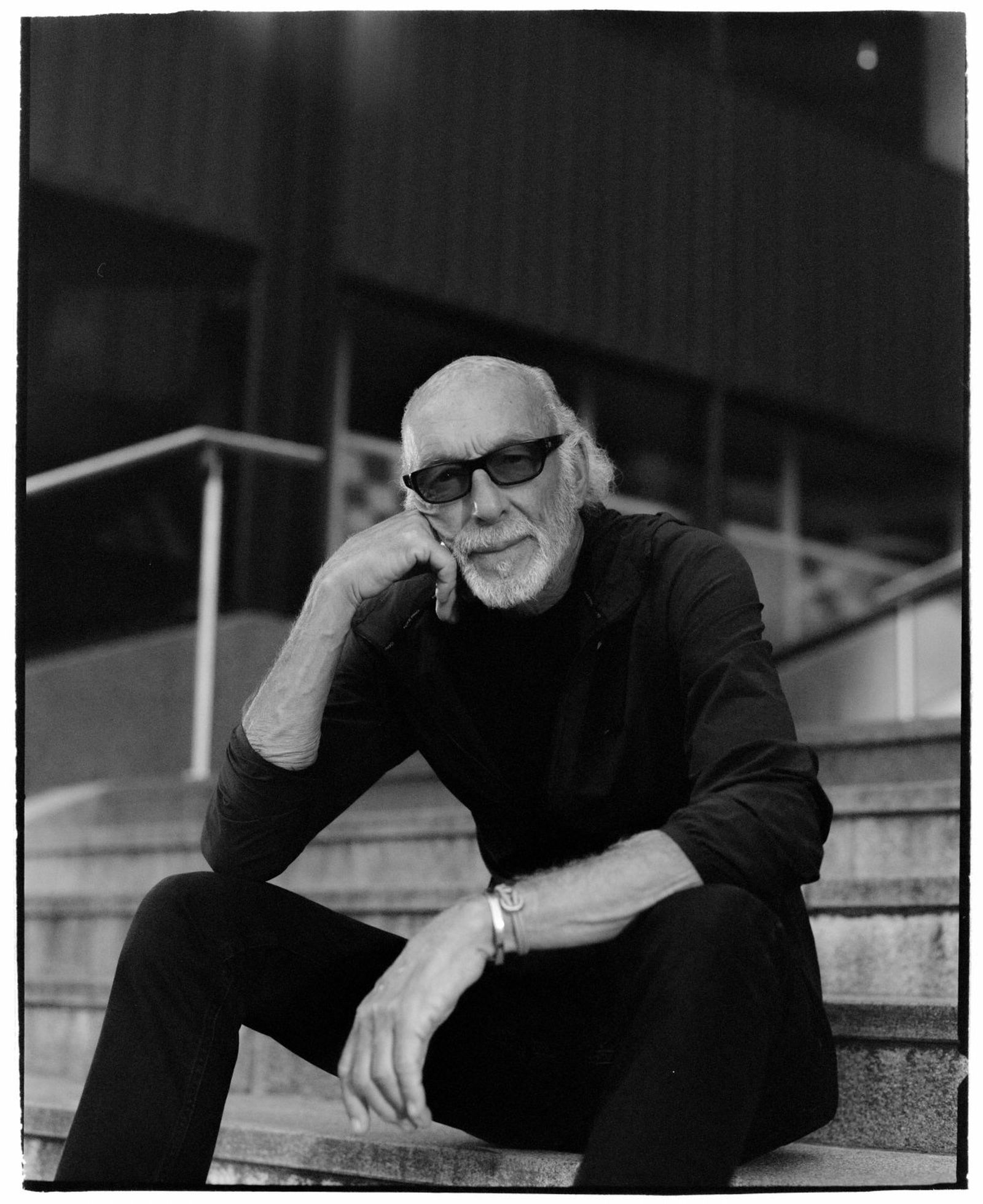 Elderly man with sunglasses and beard sitting on steps, wearing dark clothing, resting his chin on his hand, looking relaxed. Black and white photo.