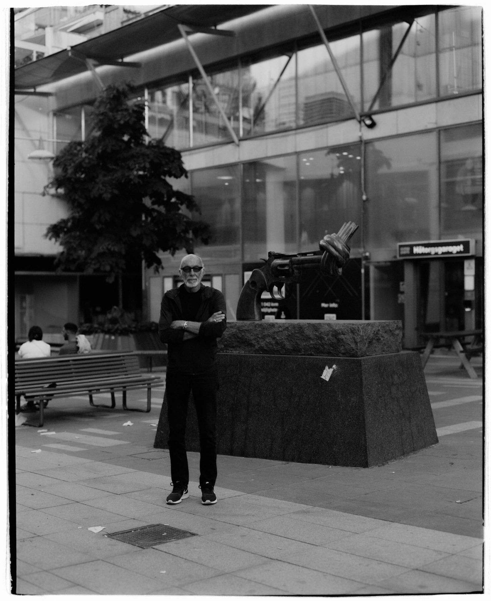 Man standing with arms crossed in front of a sculpture of a knotted gun, in an urban outdoor setting with benches and glass buildings.