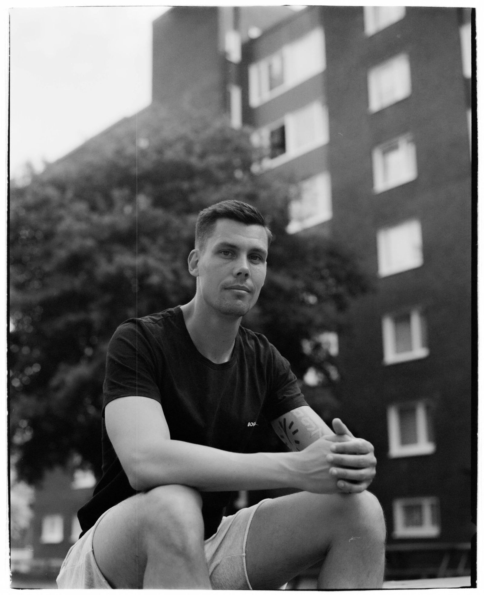 Man sitting outdoors in front of a brick building, wearing a black t-shirt and shorts, with trees in the background. Black and white photo.