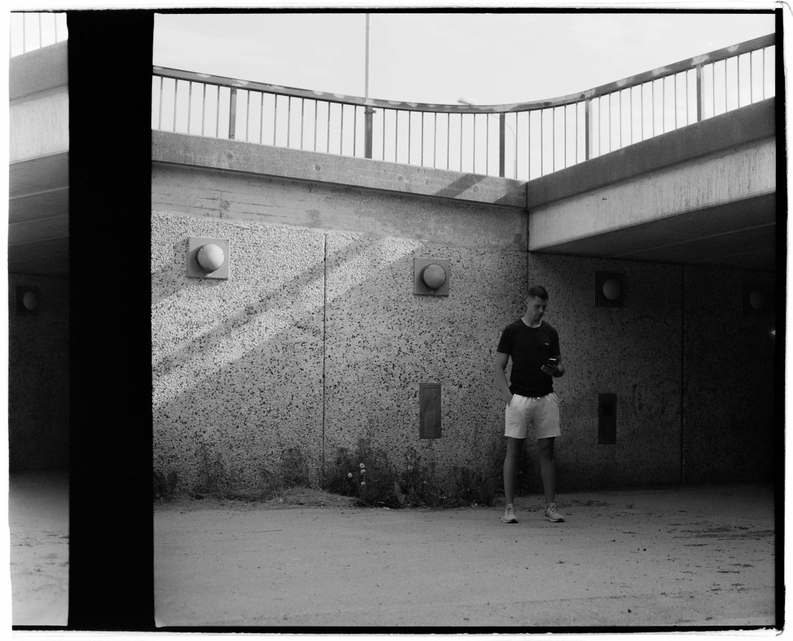 A person stands under a concrete bridge, holding an object, with sunlight casting shadows on the textured wall.