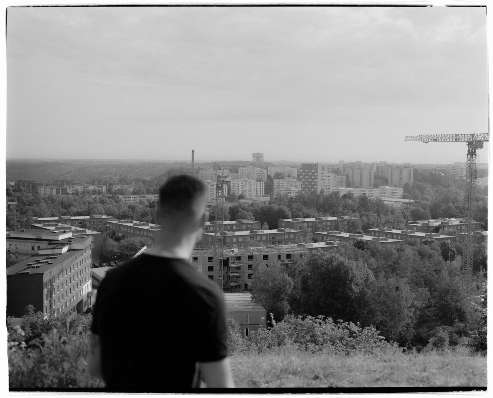 A person in a black shirt looks over a cityscape with buildings, trees, and a construction crane under a cloudy sky. Black and white photo.