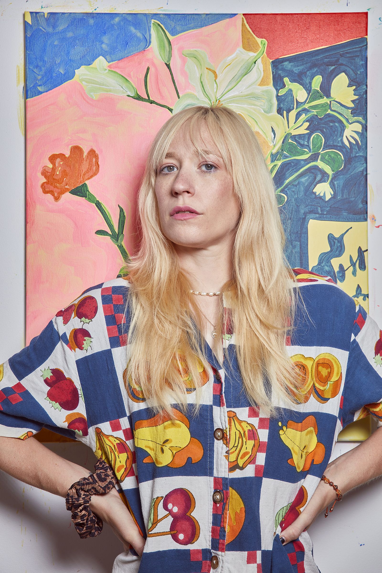 A woman with long blonde hair stands confidently in front of a colorful painting, wearing a patterned shirt with fruit designs.
