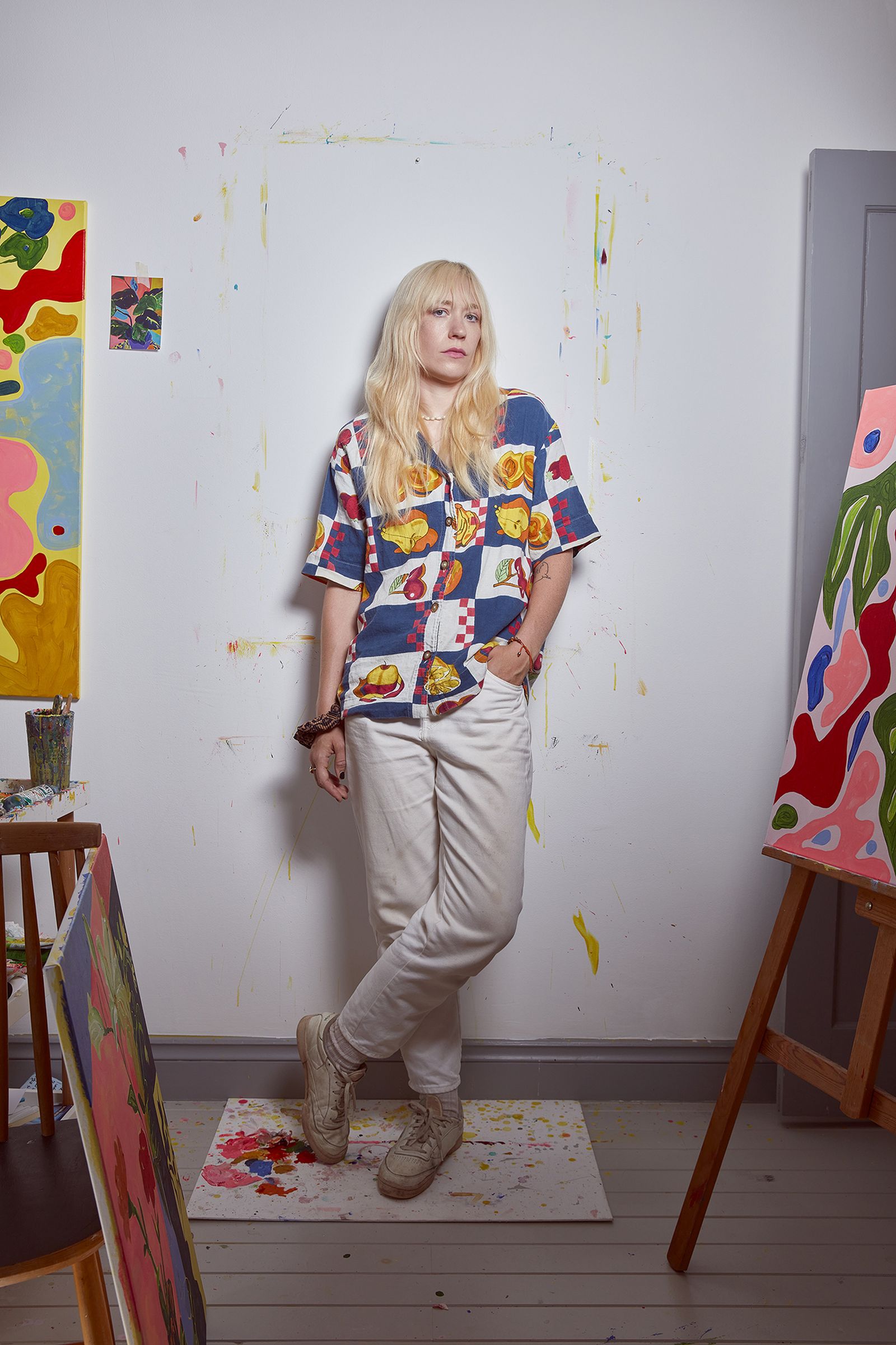 Person with long blonde hair in a colorful shirt stands in an art studio surrounded by vibrant abstract paintings on easels.