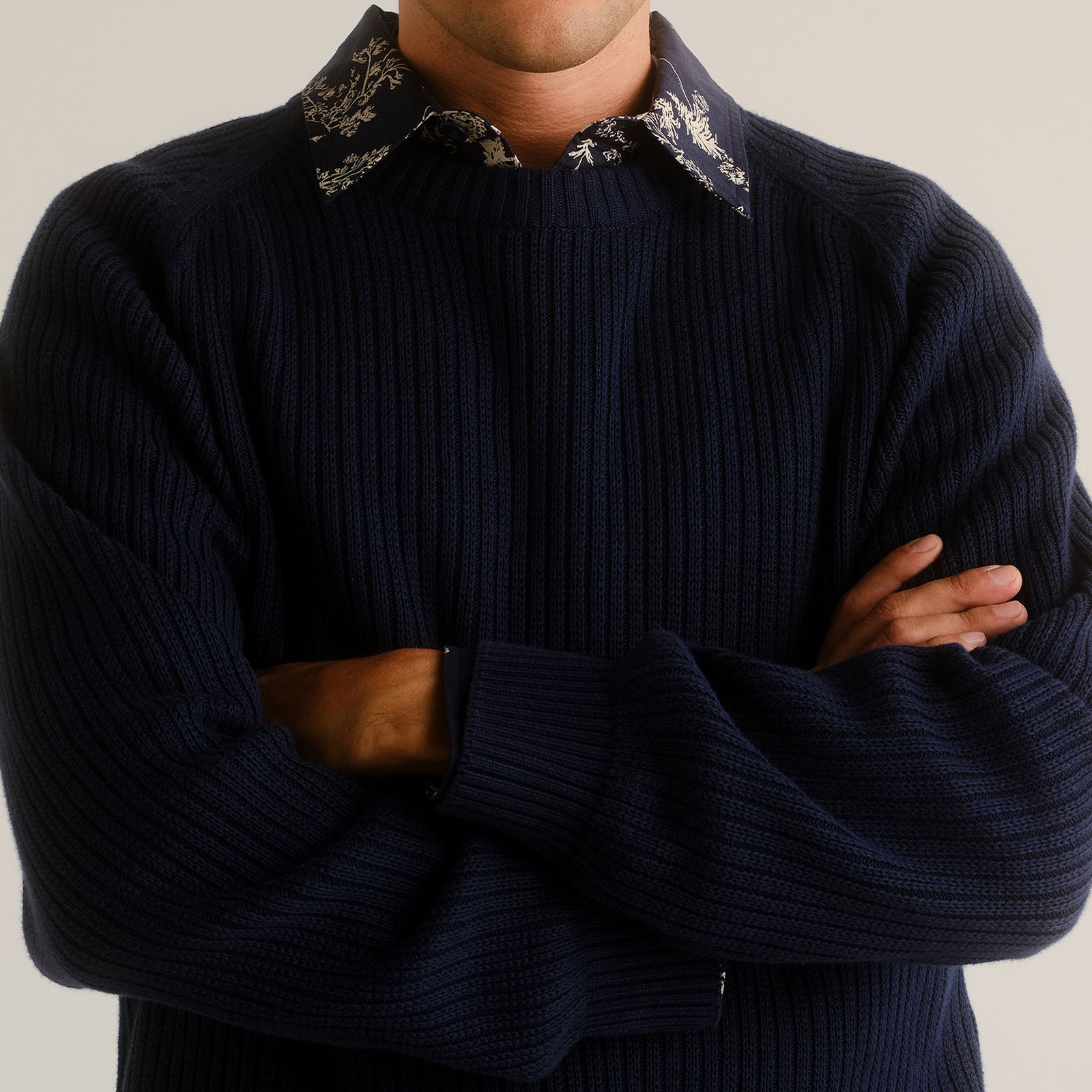 Man in a dark ribbed sweater and patterned collar shirt, arms crossed, standing against a neutral background.