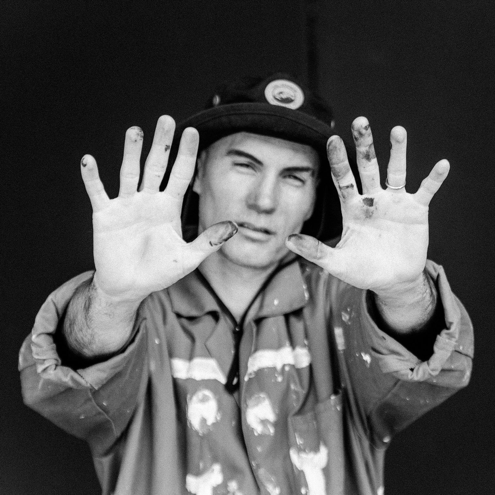 A person in a work jacket and cap holds up hands with paint stains, against a dark background, in a black and white image.
