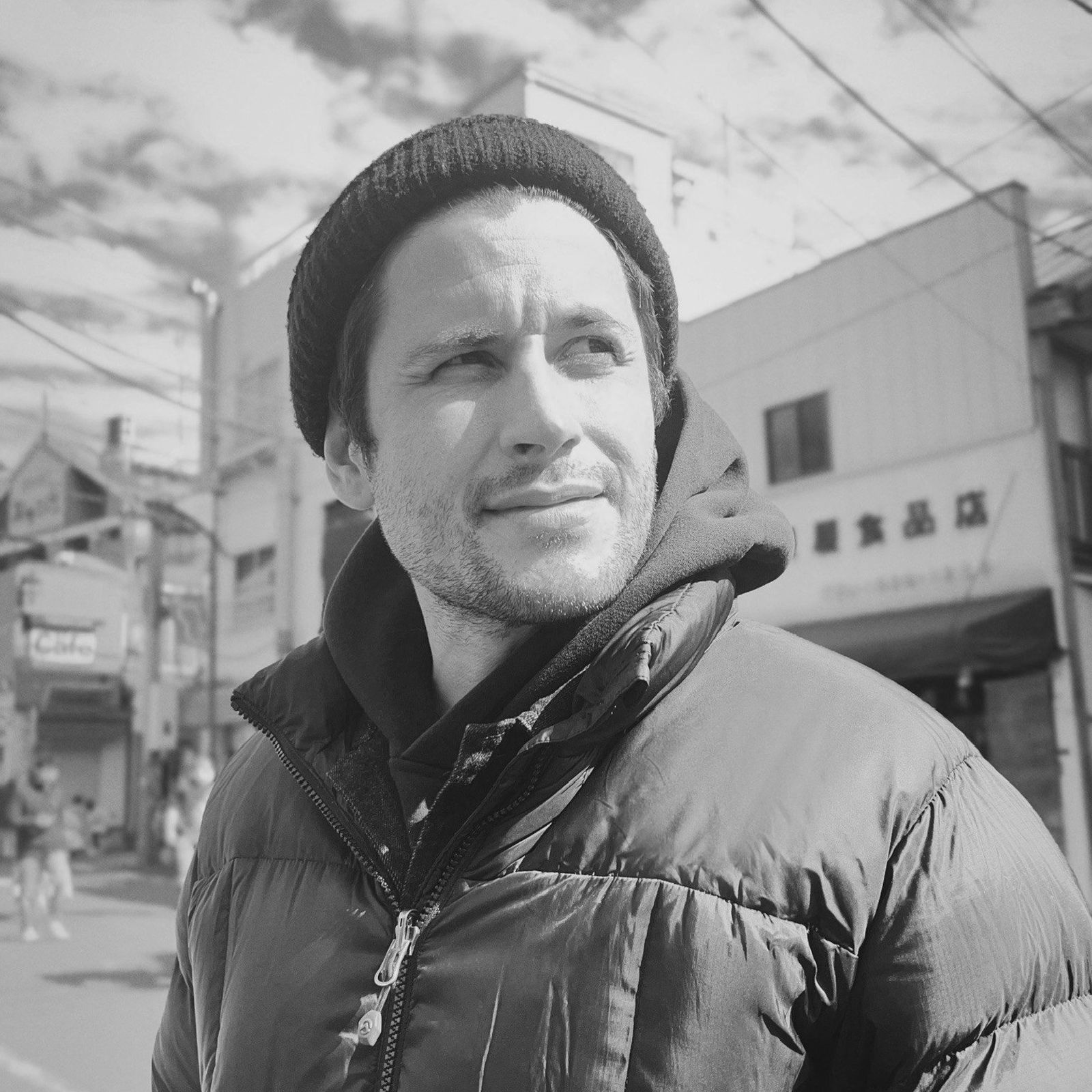 A person in a beanie and puffer jacket looks to the side on a street with buildings and power lines in the background. Black and white photo.