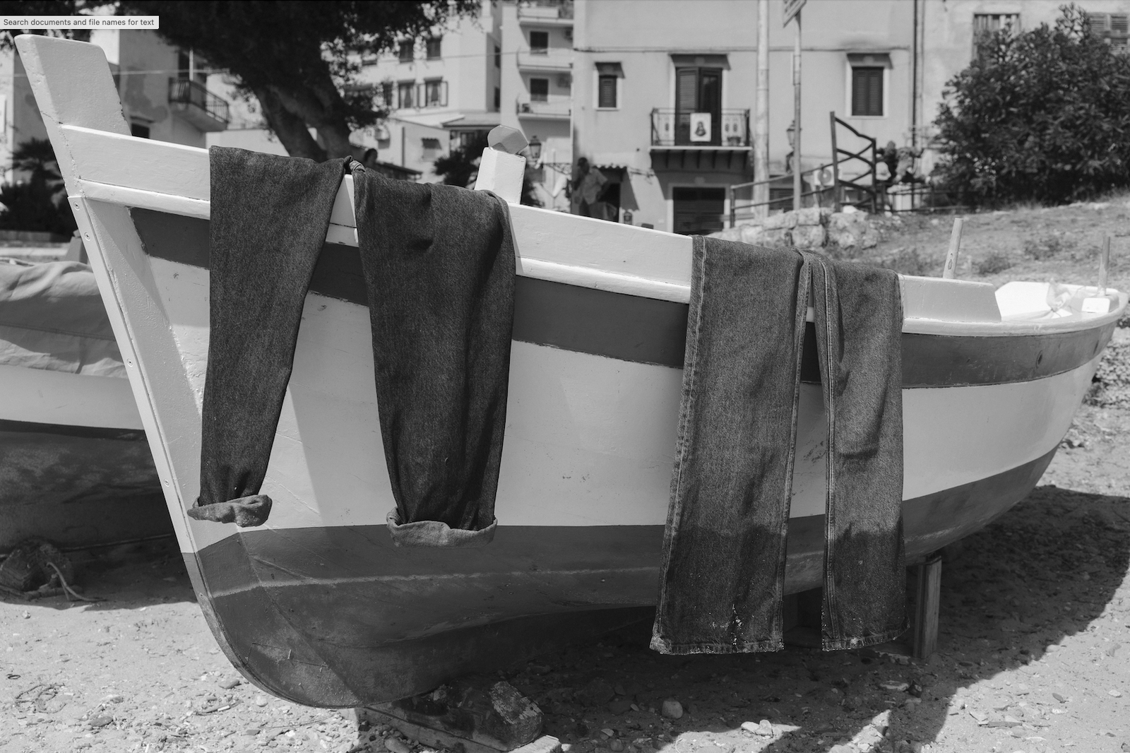 Three pairs of blue jeans draped over a small, docked boat on a sandy beach, with buildings and foliage in the background.