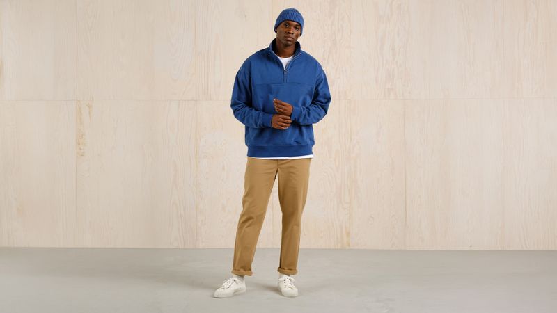 Man in a blue sweater and beanie, beige pants, and white sneakers stands against a light wood background.