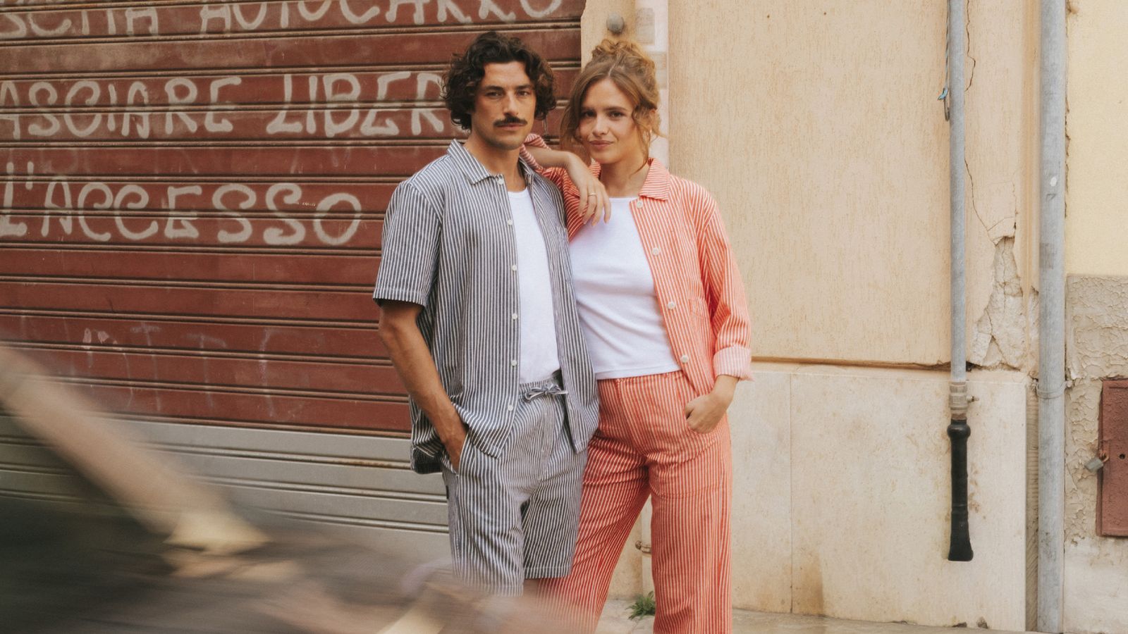 Two persons standing infront of a car thats swoshing by. One guy with mustash and curly hair standing with his hand in a pocket with a striped blue and white short shirt and matching shorts. A girl is leaning on his sholder with a striped red and white jacket and matching pants.