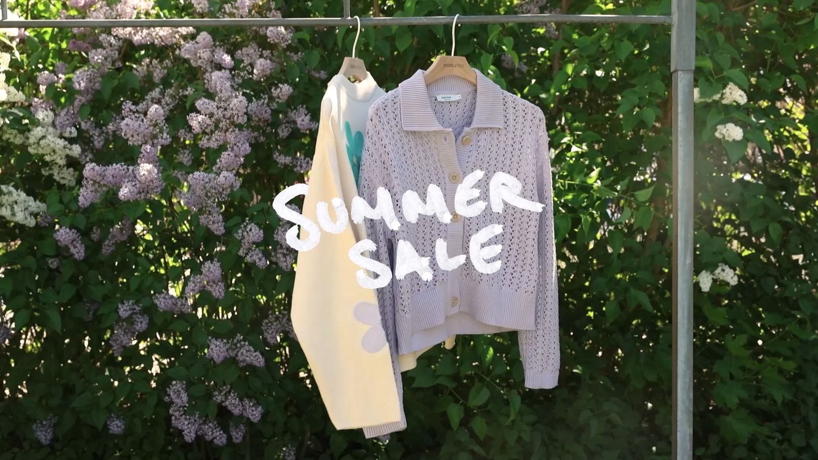 Two sweaters on hangers with "Summer Sale" text, set against a backdrop of lush green foliage and blooming lilac flowers.