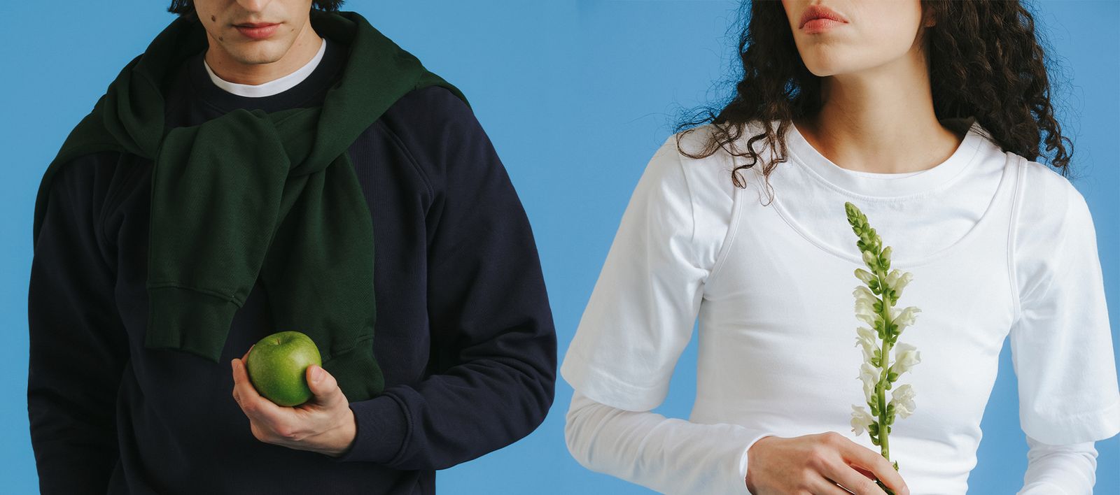 Two people against a blue background; one holds a green apple wearing a dark sweater, the other holds a white flower wearing a white top.