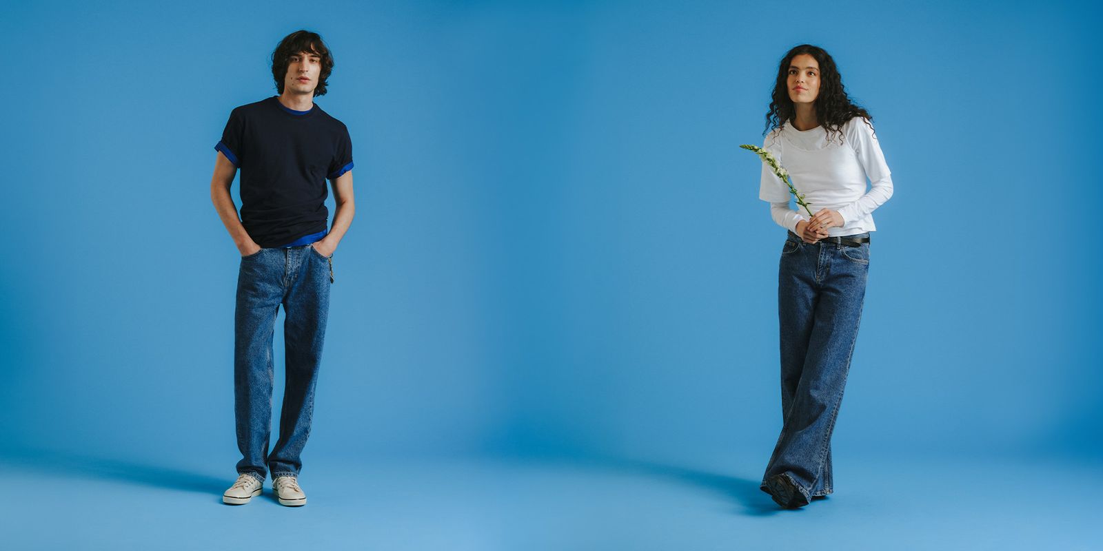 Two people stand against a blue background; one in a black shirt and jeans, the other in a white shirt and jeans holding a flower.