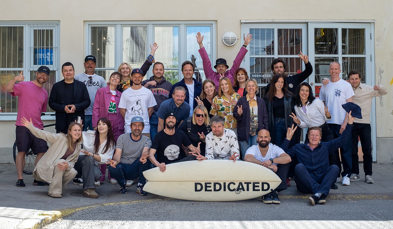 A diverse group of people posing cheerfully outside a building, holding a surfboard with "DEDICATED" written on it.