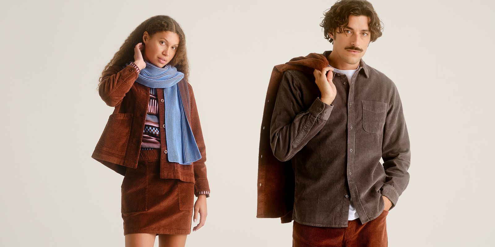 A woman in a brown outfit with a blue scarf and a man in a brown jacket with a gray shirt pose against a neutral background.