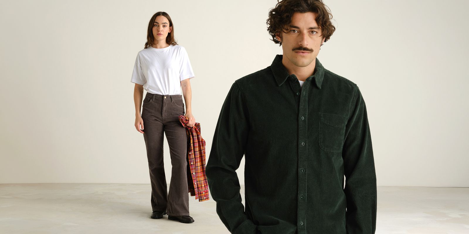 A man in a dark green corduroy shirt stands in the foreground, while a woman in a white tee and dark pants holds a plaid shirt in the background.