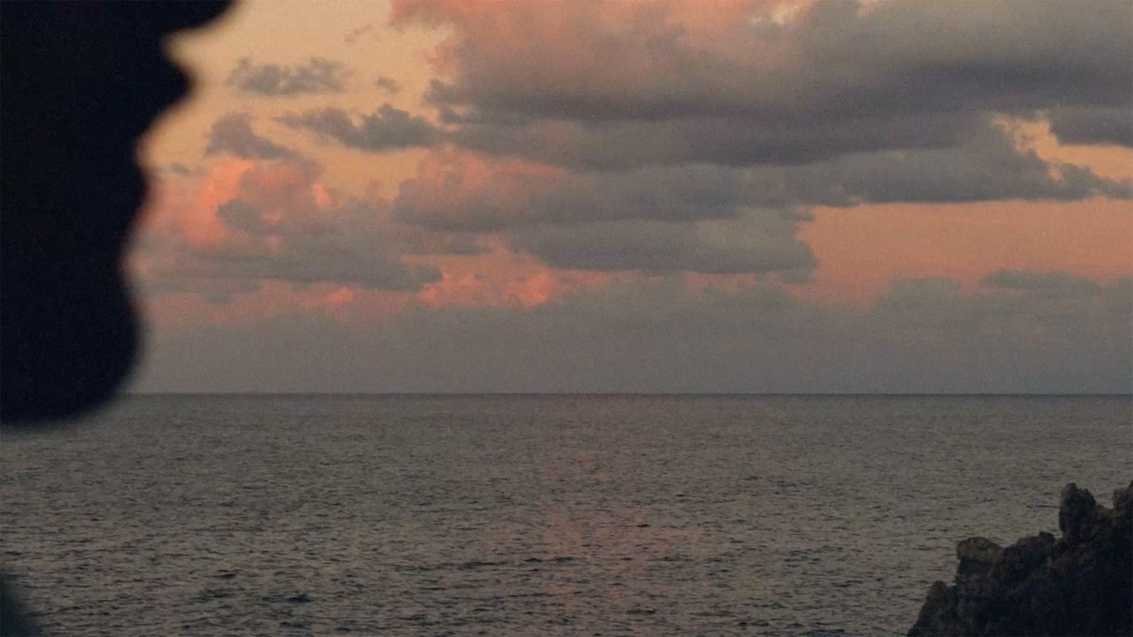 Silhouette of a person against a serene ocean backdrop at sunset, with pink-tinged clouds scattered across the sky.