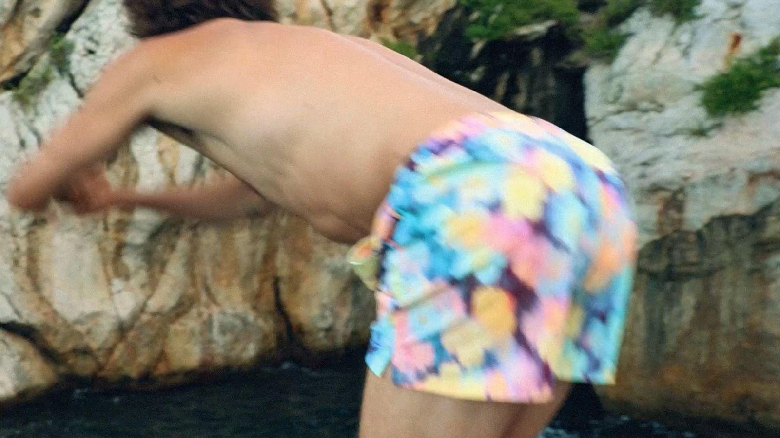 Person wearing colorful shorts, bending over by rocky cliffs with greenery nearby.