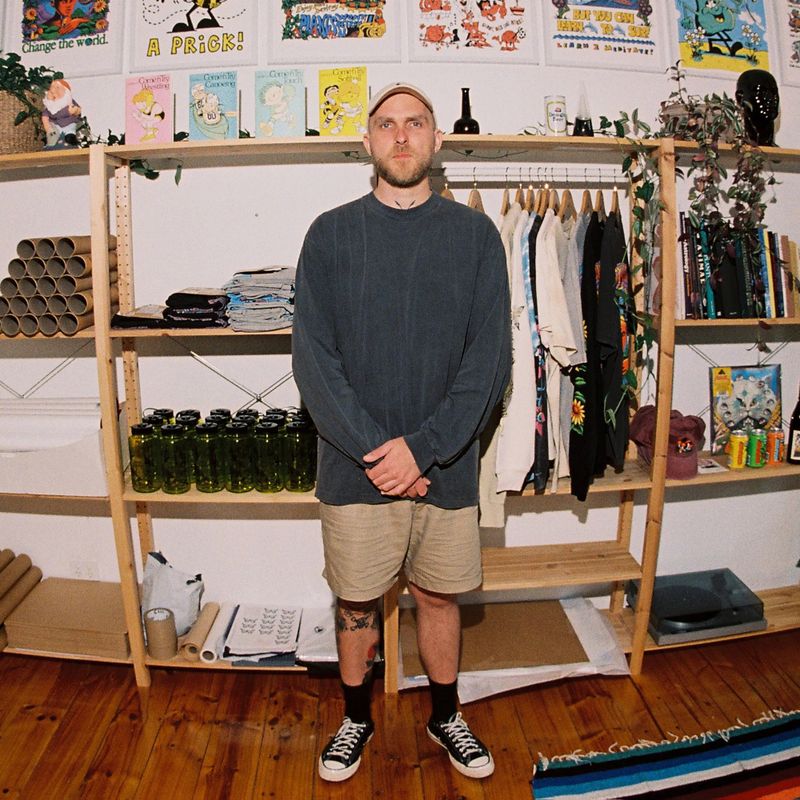 Man in casual clothing stands in front of a wooden shelf with clothes, posters, and green bottles in a room with wooden flooring.