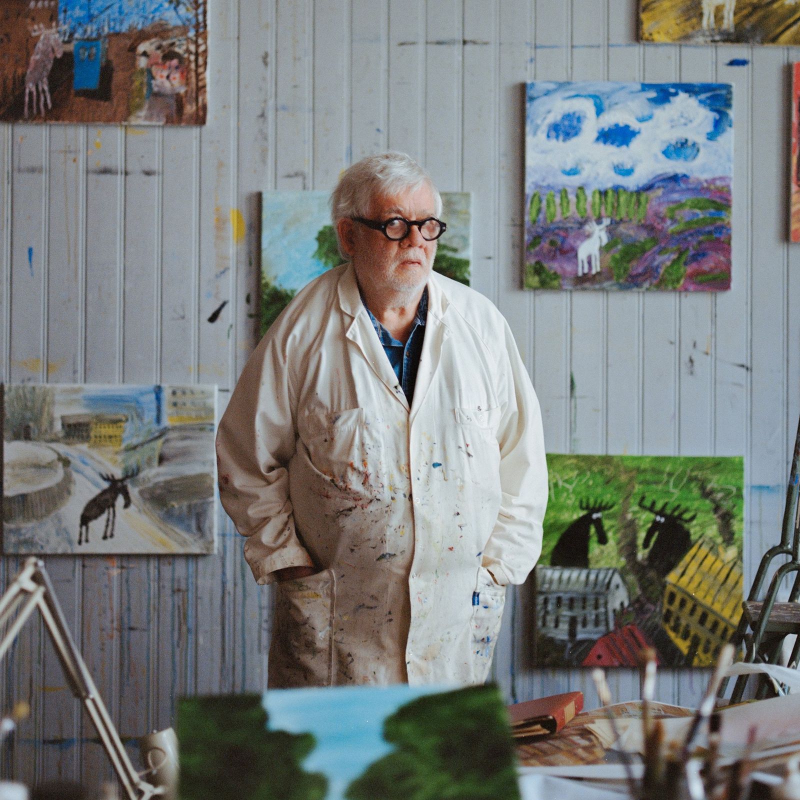 Elderly man in a paint-splattered smock stands in an art studio, surrounded by colorful landscape paintings on the walls.