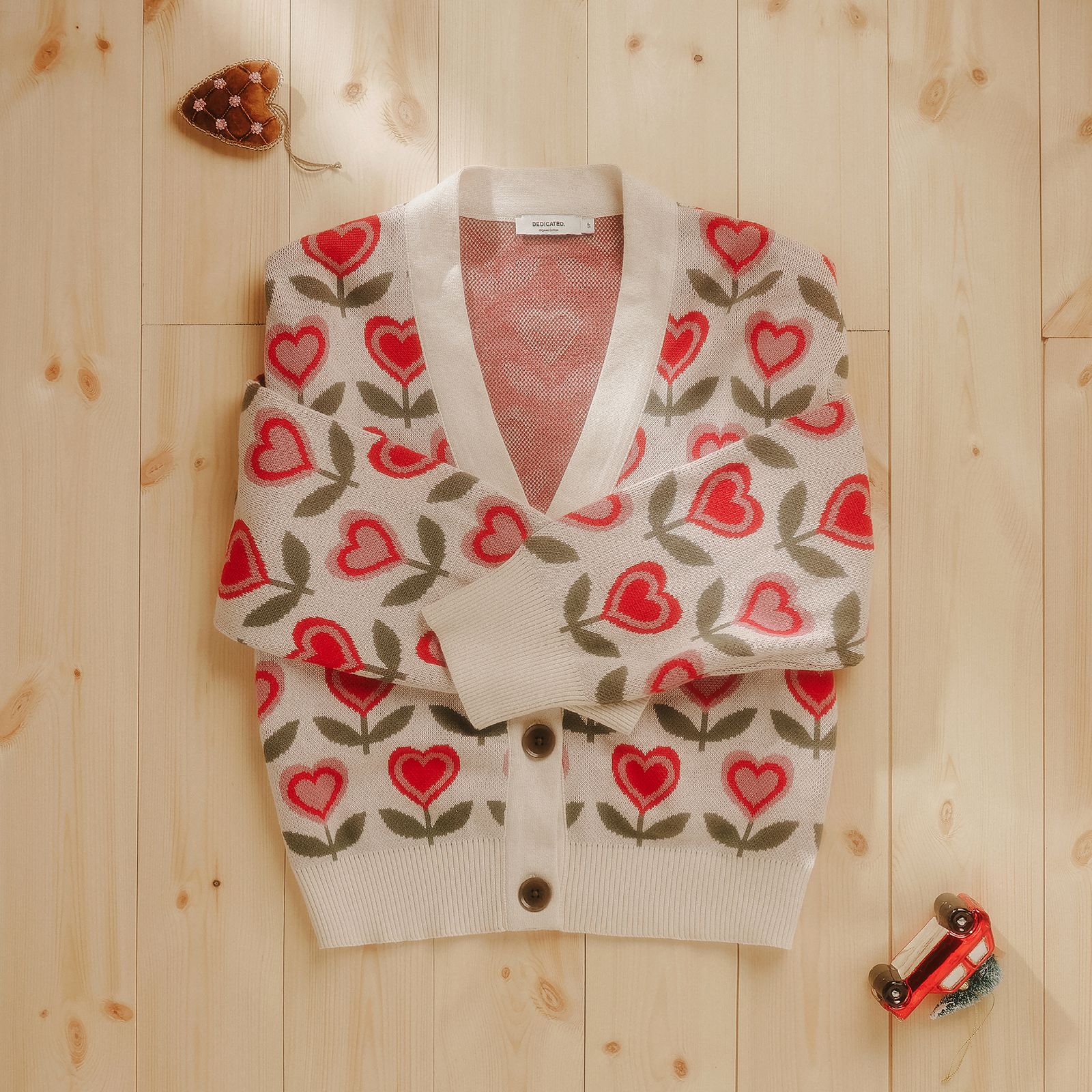 Cream cardigan with red heart and green leaf pattern on wooden floor, surrounded by a small toy car and a heart-shaped decoration.