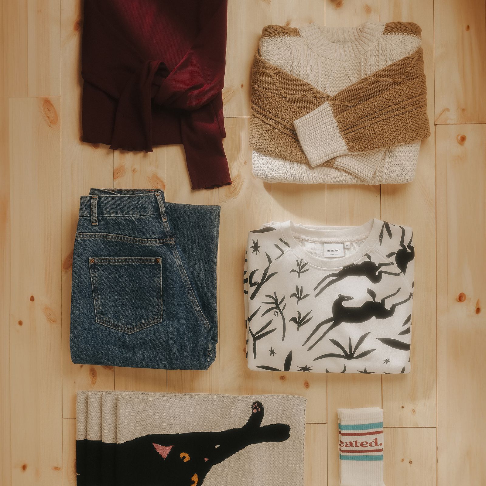 Folded clothes on a wooden floor: sweaters, jeans, a graphic tee, a scarf with a black cat design, and striped socks.