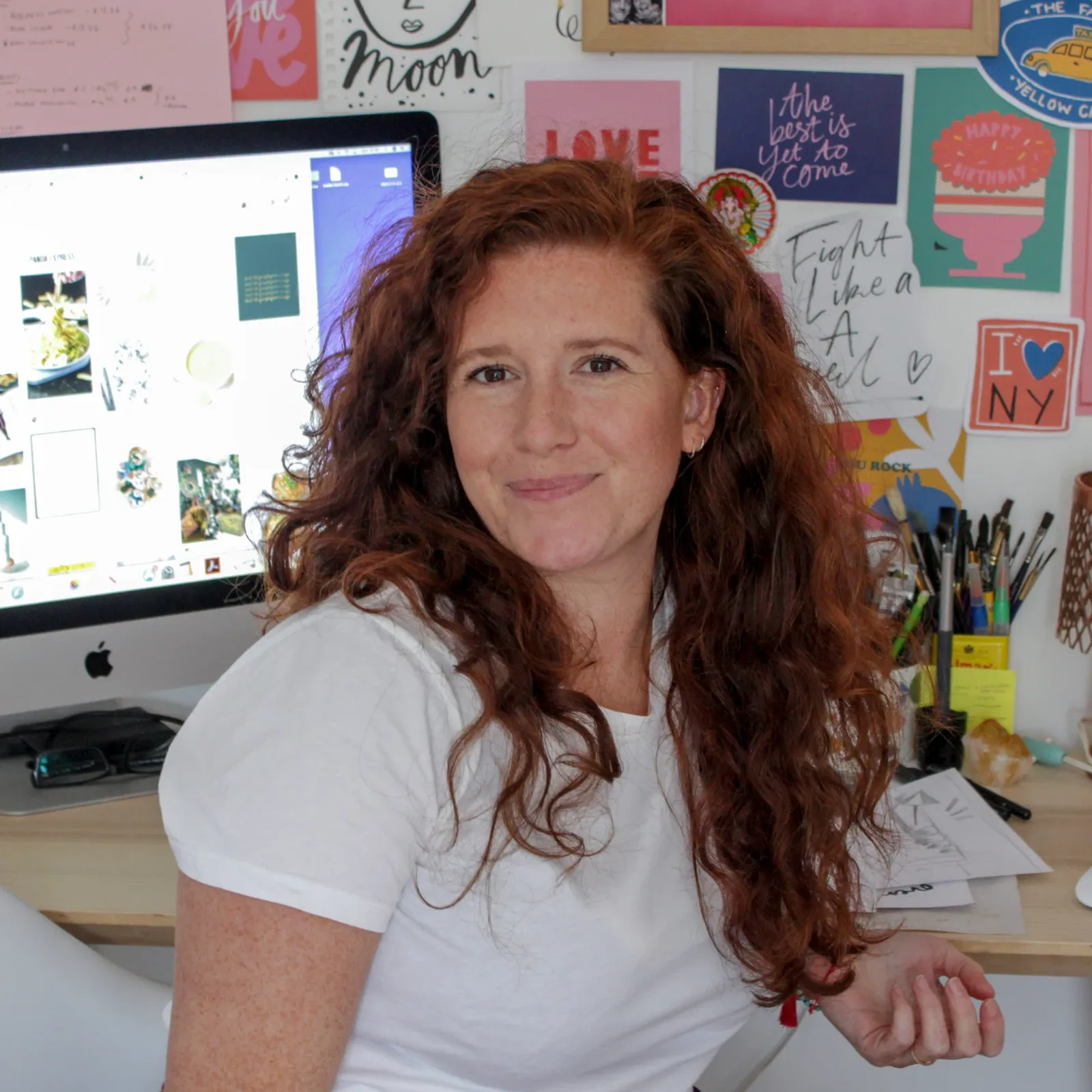 Person with long red hair smiling at a desk, surrounded by colorful wall art and a computer displaying various images.