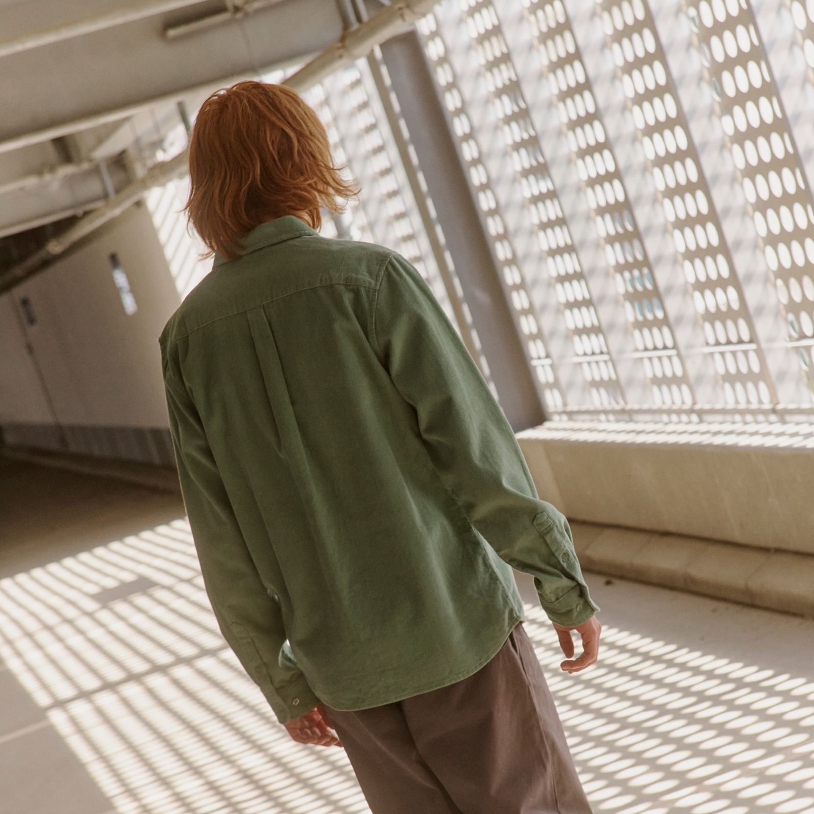 Person with shoulder-length hair in a green shirt walks down a sunlit corridor with patterned shadows on the floor and walls.