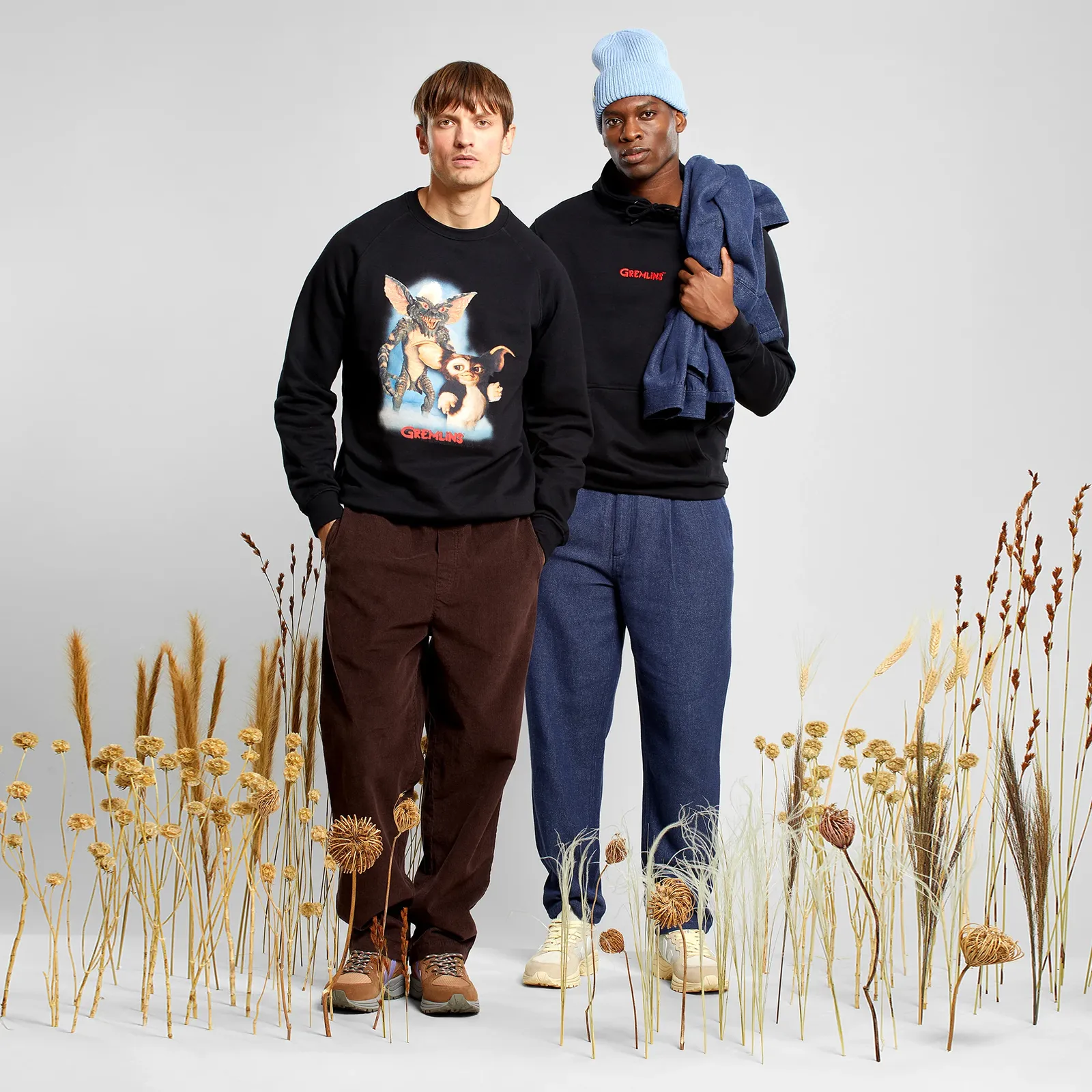 Two men in casual outfits stand among dried plants. One wears a black sweater with a graphic, and the other a blue beanie and holding a jacket.