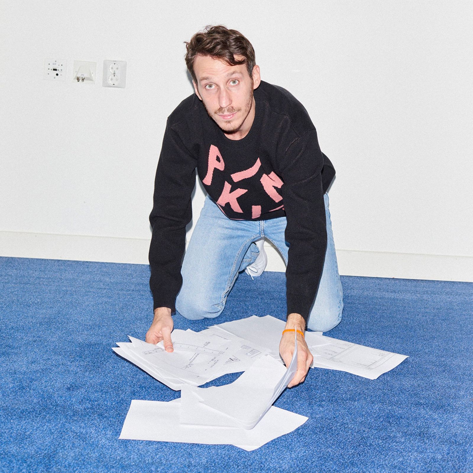 A person kneels on a blue carpet, gathering scattered papers. They wear a black sweater with pink letters and light blue jeans.