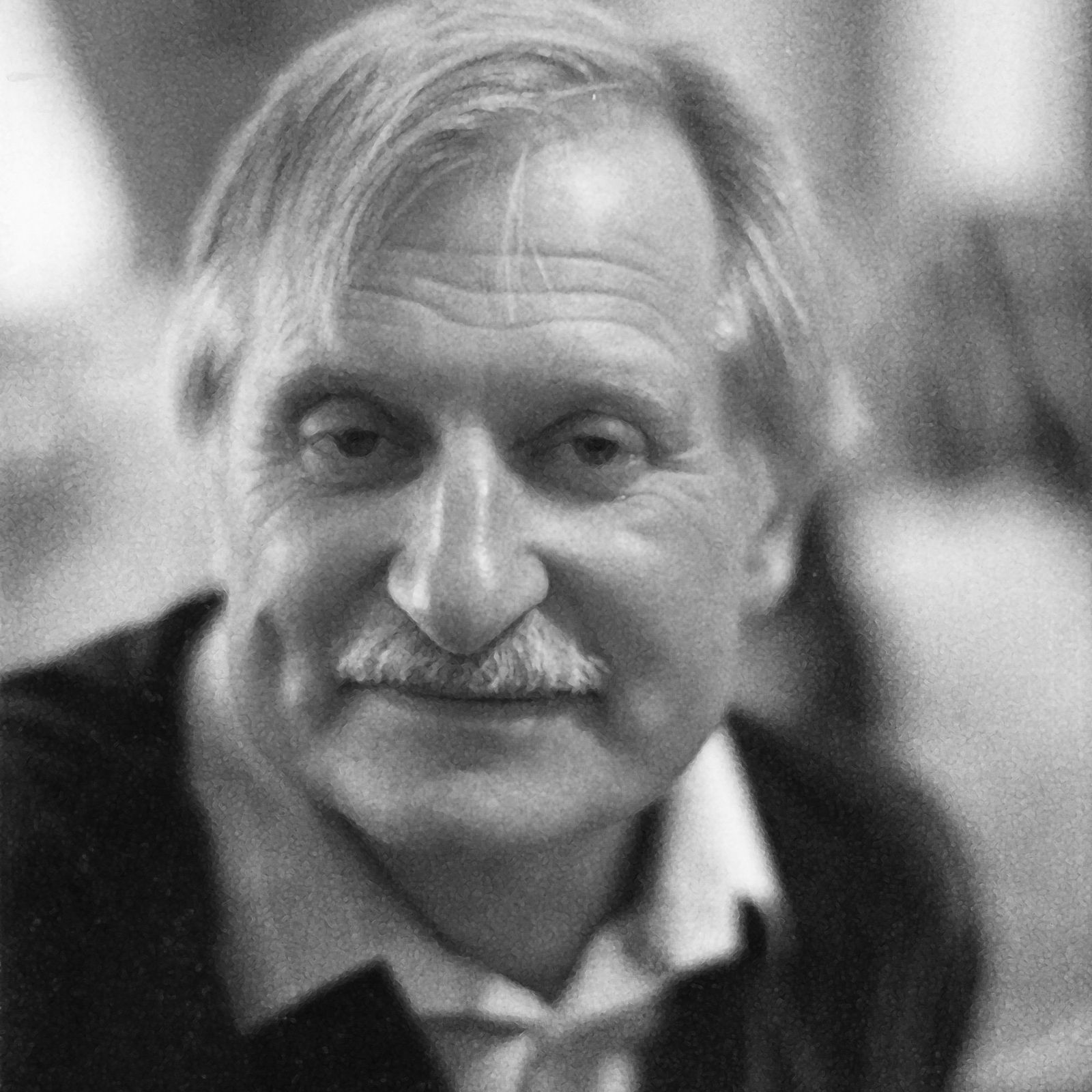 Black and white portrait of an older man with light hair and a mustache, wearing a collared shirt and sweater, looking into the camera.