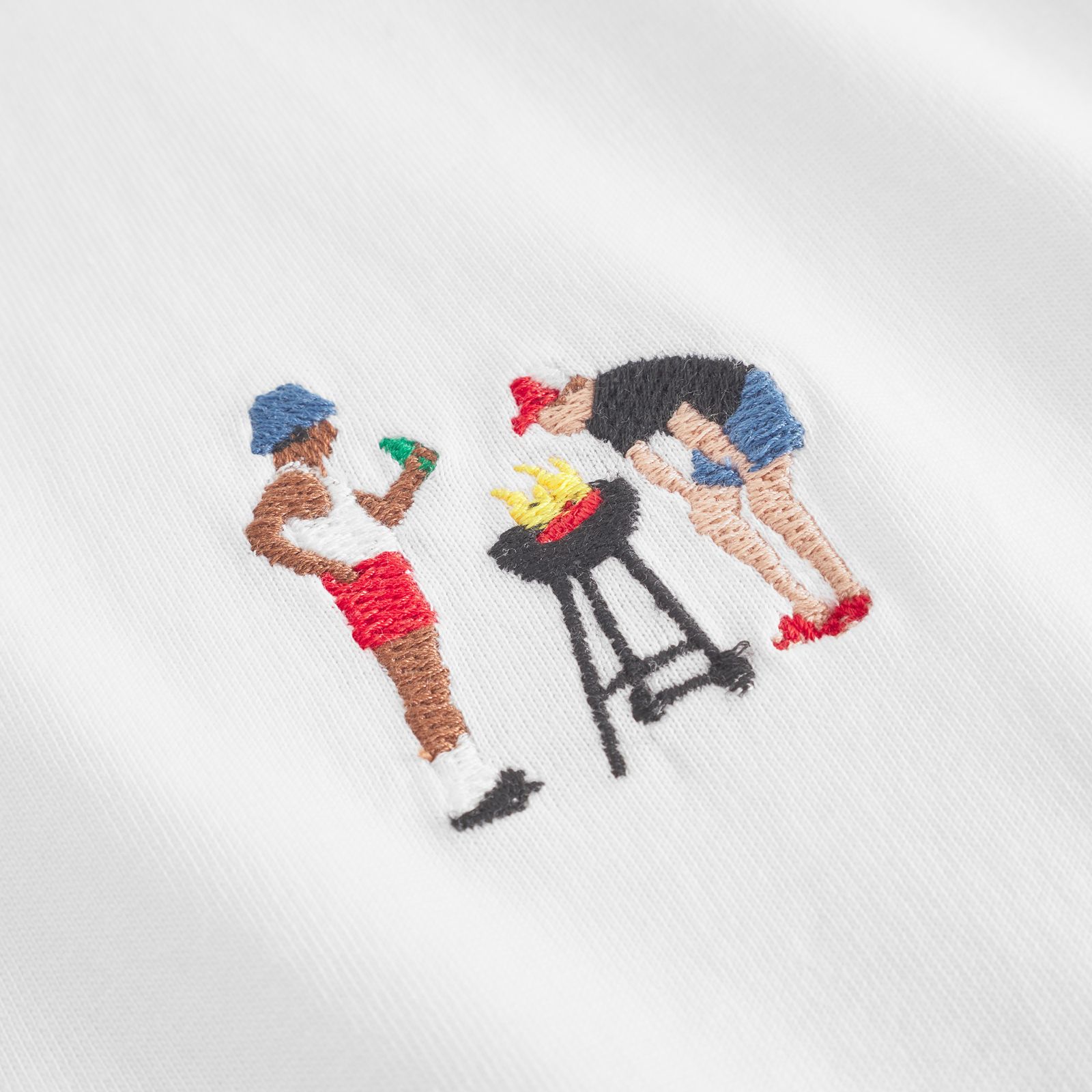 Embroidery of two people grilling; one holds a drink, the other tends to a barbecue. Both wear colorful shorts and hats on a white fabric.