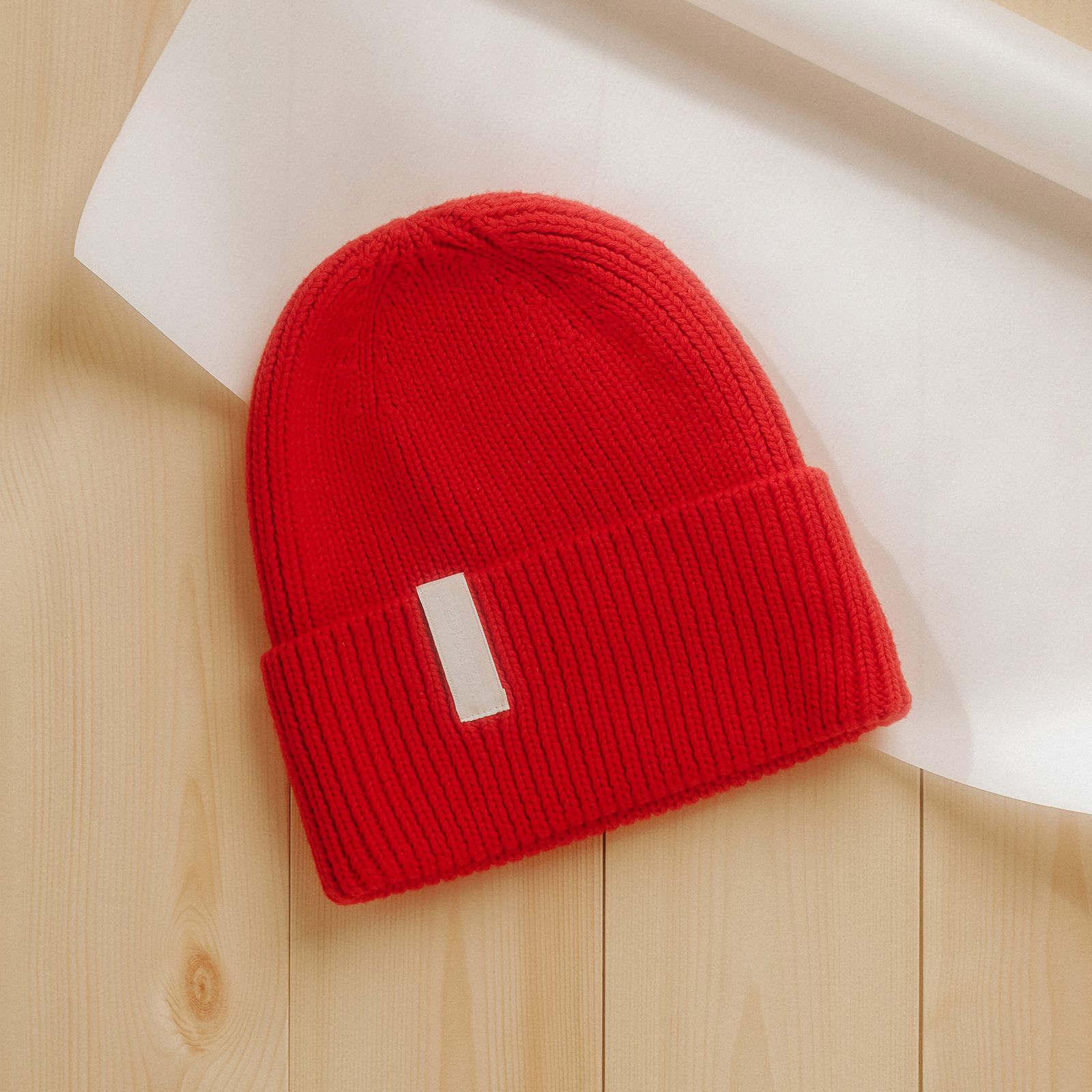 Red knit beanie with a white label, placed on light wooden surface with a partially unrolled sheet of white paper.